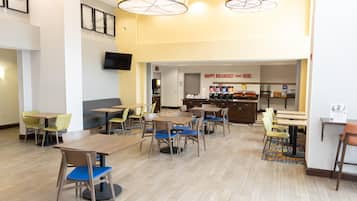 A hotel dining area with multiple tables and chairs, a television mounted on the wall, and a breakfast counter with coffee options.