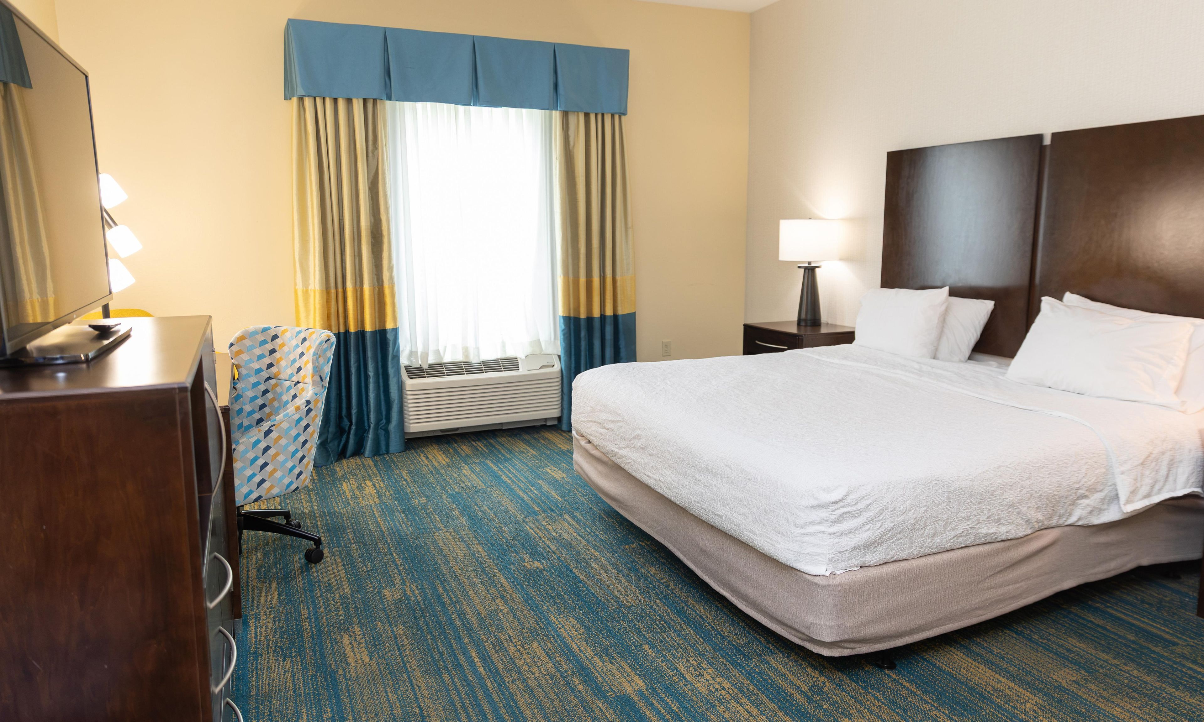 Quarto de hotel com cama, escrivaninha, cadeira, abajur e janela com cortinas.