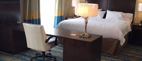 Une chambre d’hôtel équipée d’un lit, d’un bureau, d’une chaise, d’une lampe et d’une grande fenêtre avec des rideaux.
