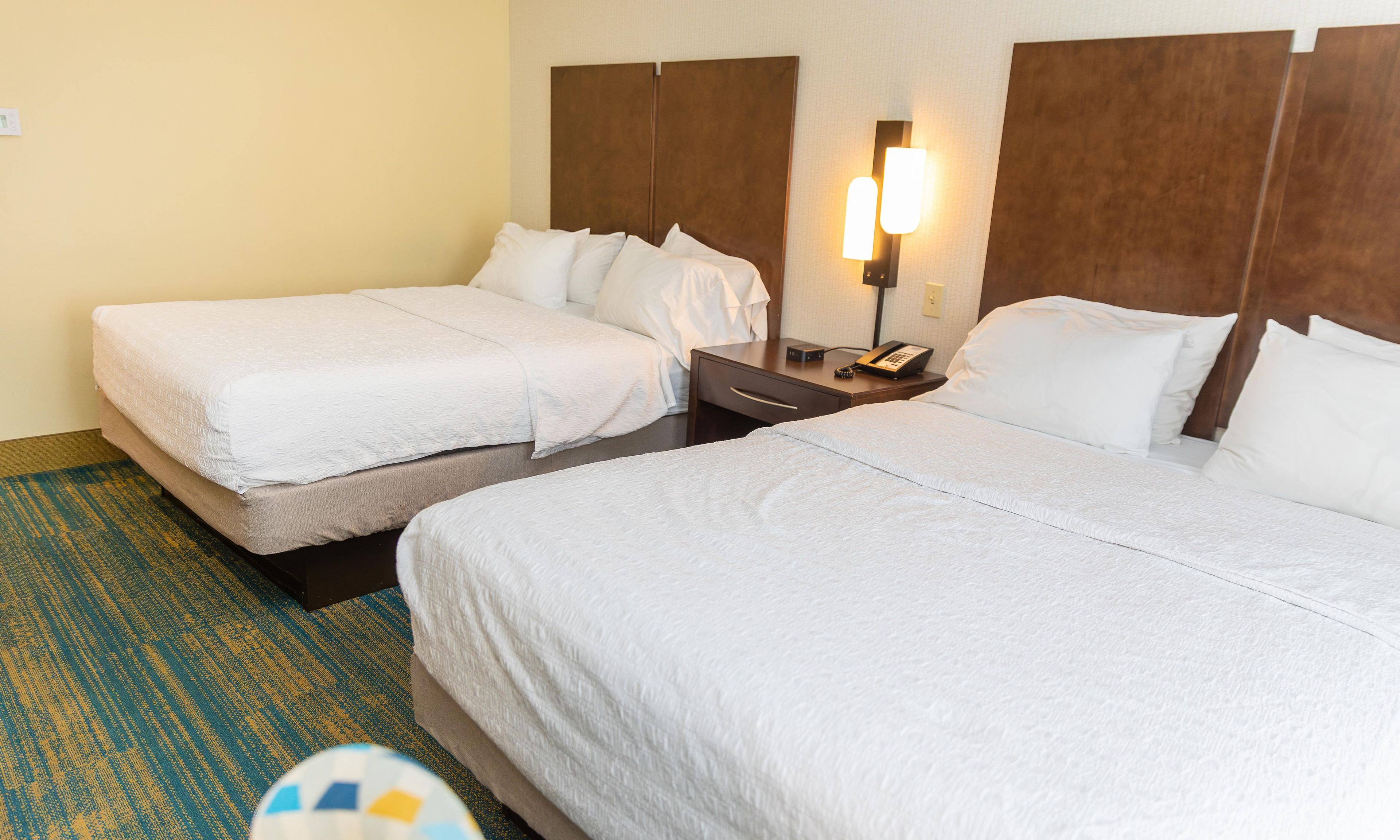 A hotel room with two beds, a nightstand with a phone, and a wall-mounted light.