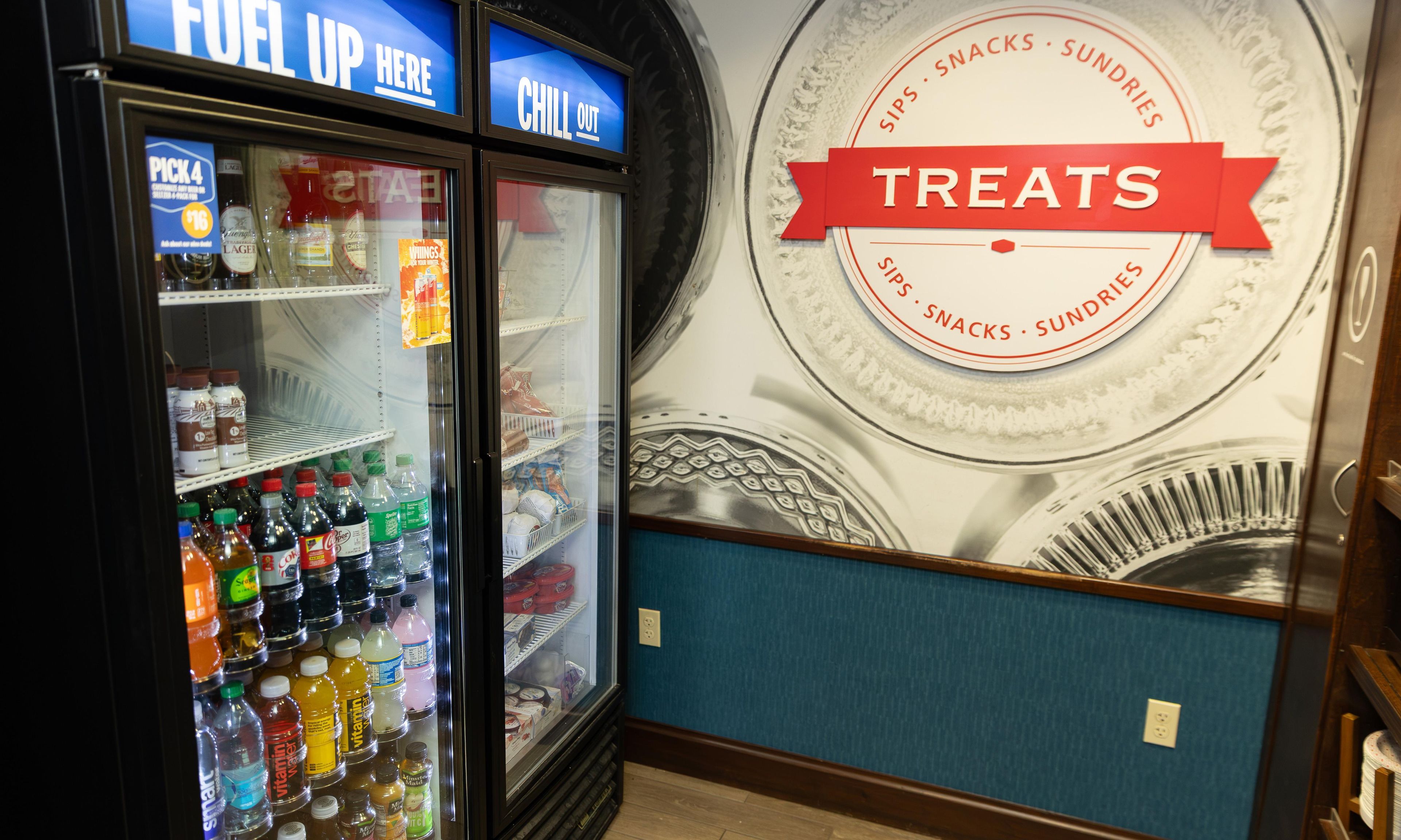 A beverage cooler stocked with various drinks and a wall sign advertising treats.