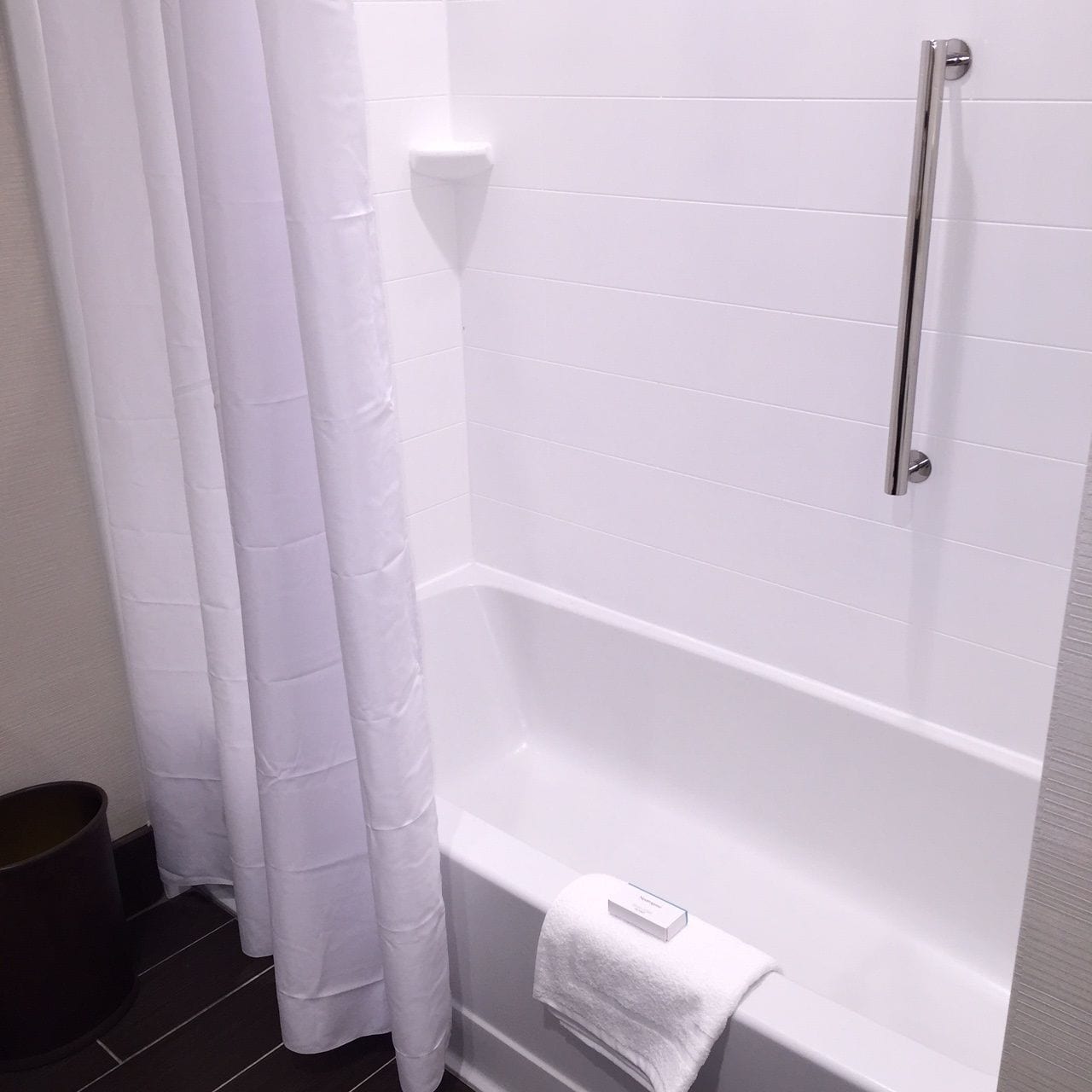 A white bathroom with a bathtub, a handrail, and a white towel on the edge.