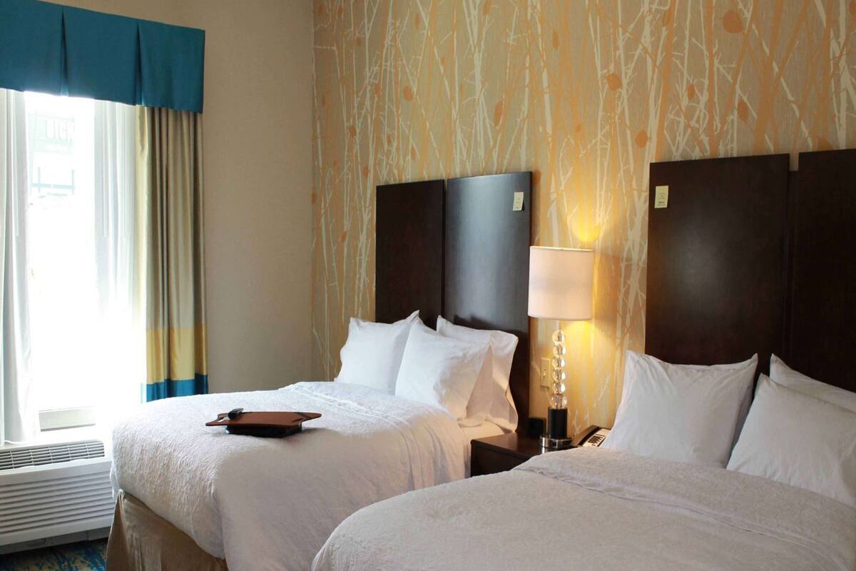 Habitación de hotel con dos camas, un ventanal con cortinas, pared con diseño y suelo alfombrado.