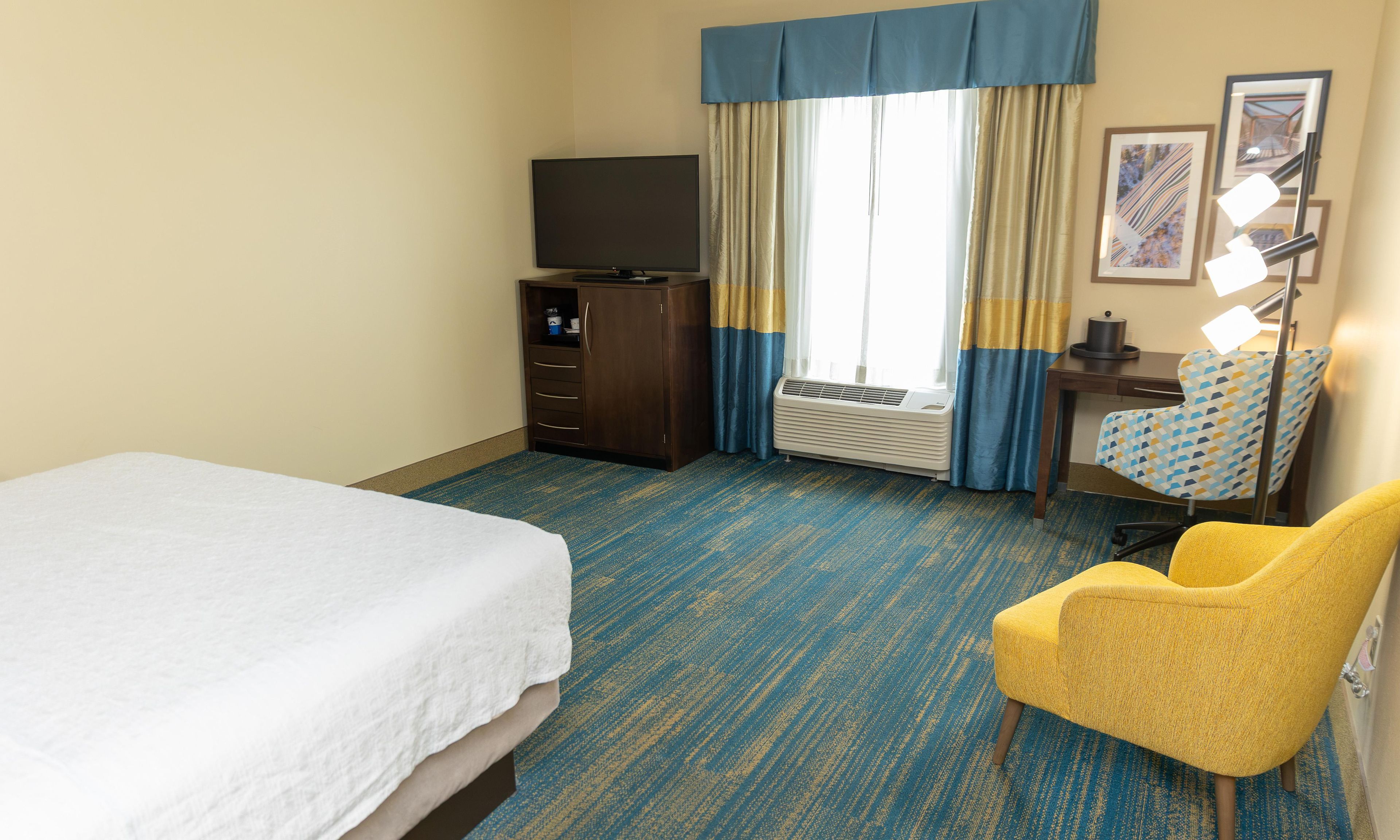 Habitación de hotel con cama, escritorio, silla, televisor y ventana con cortinas.