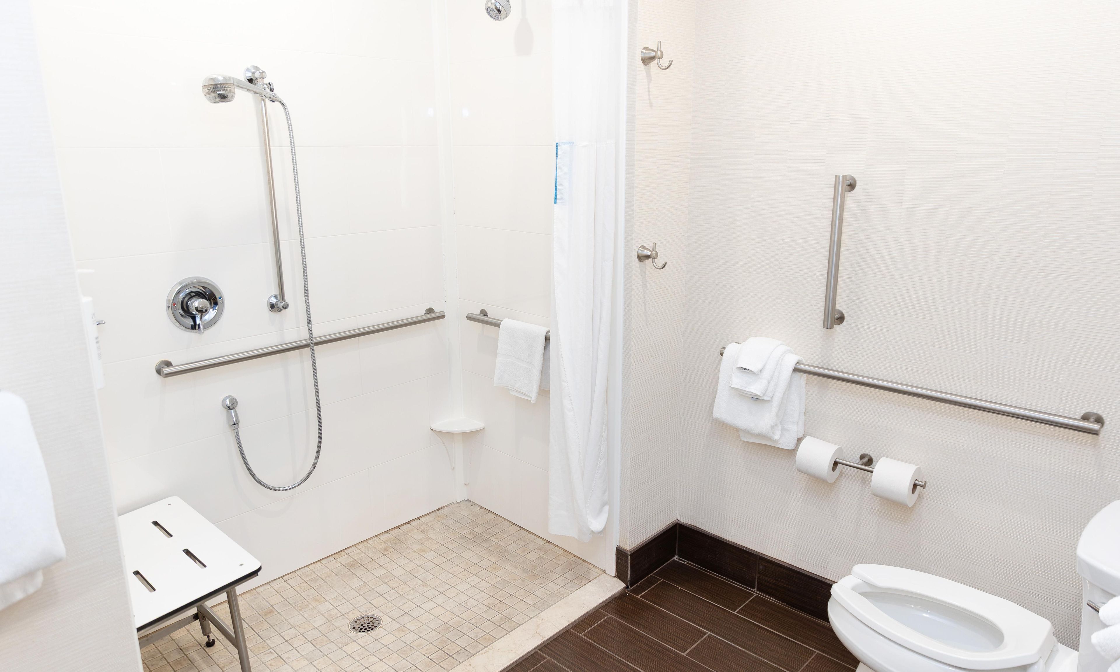 Une salle de bain équipée d’une douche, de toilettes, de barres d’appui et d’un banc accessible aux personnes en fauteuil roulant.