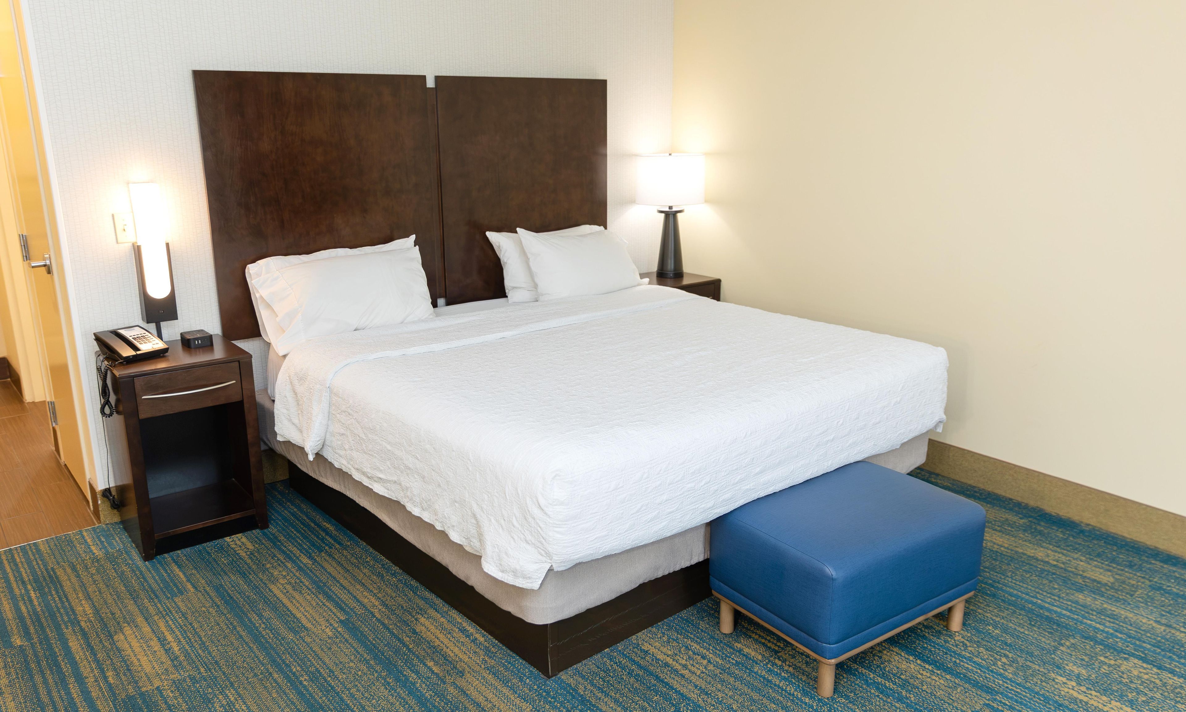 A hotel room with a large bed, bedside lamps, a phone on the nightstand, and a blue bench.