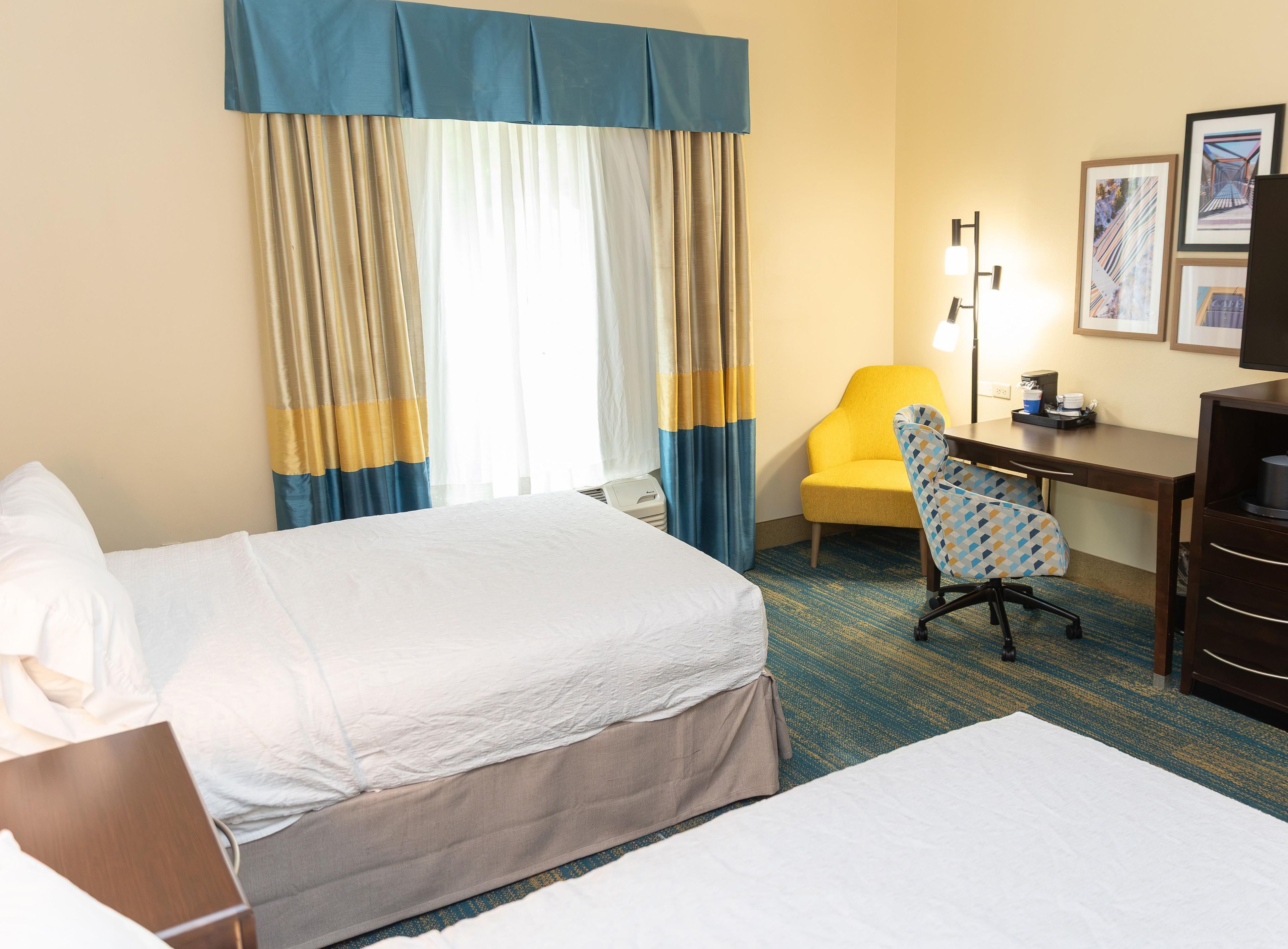 Quarto de hotel com duas camas, uma escrivaninha, uma cadeira e um abajur.