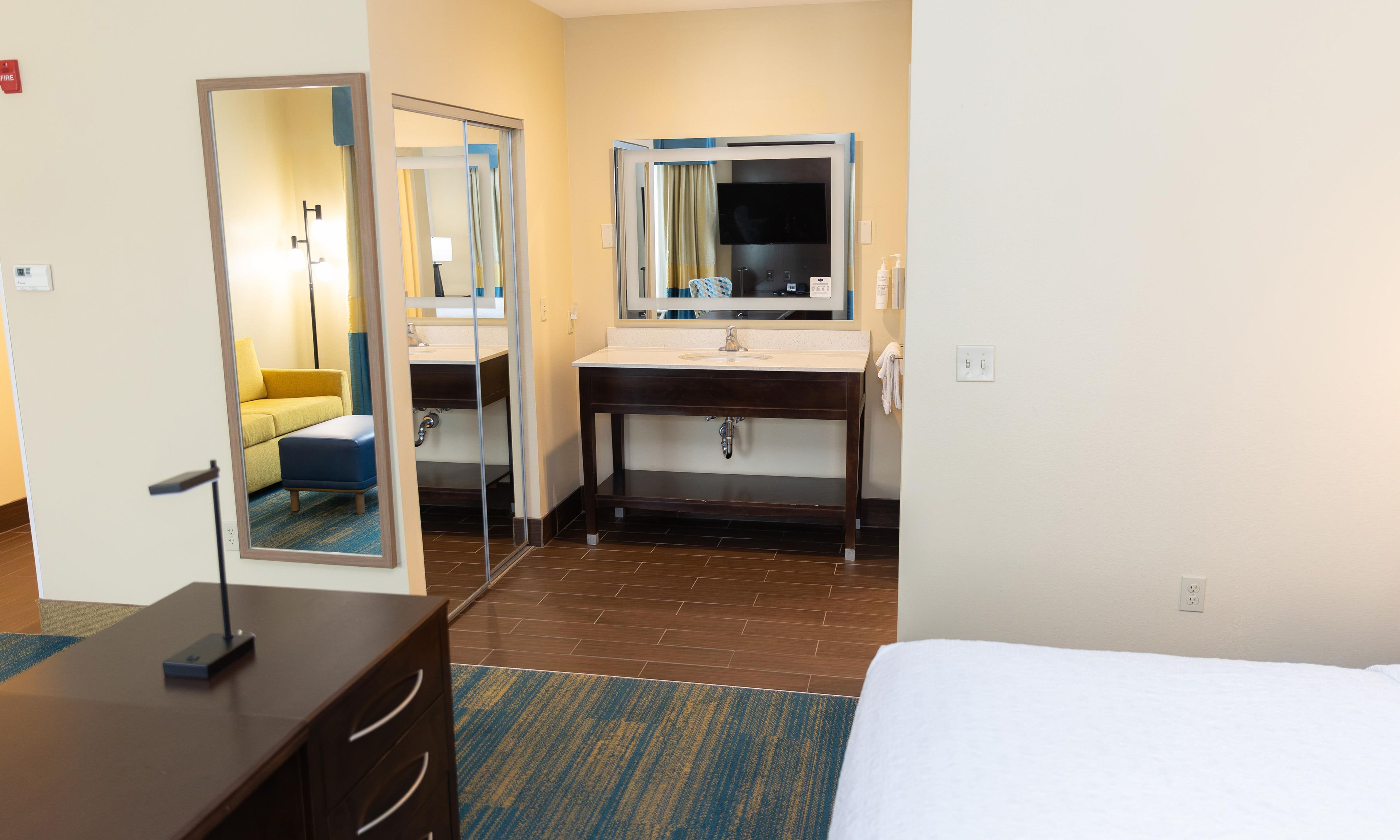 A hotel room with a bed, a wooden dresser, a bathroom with a mirror, sink, and a television.