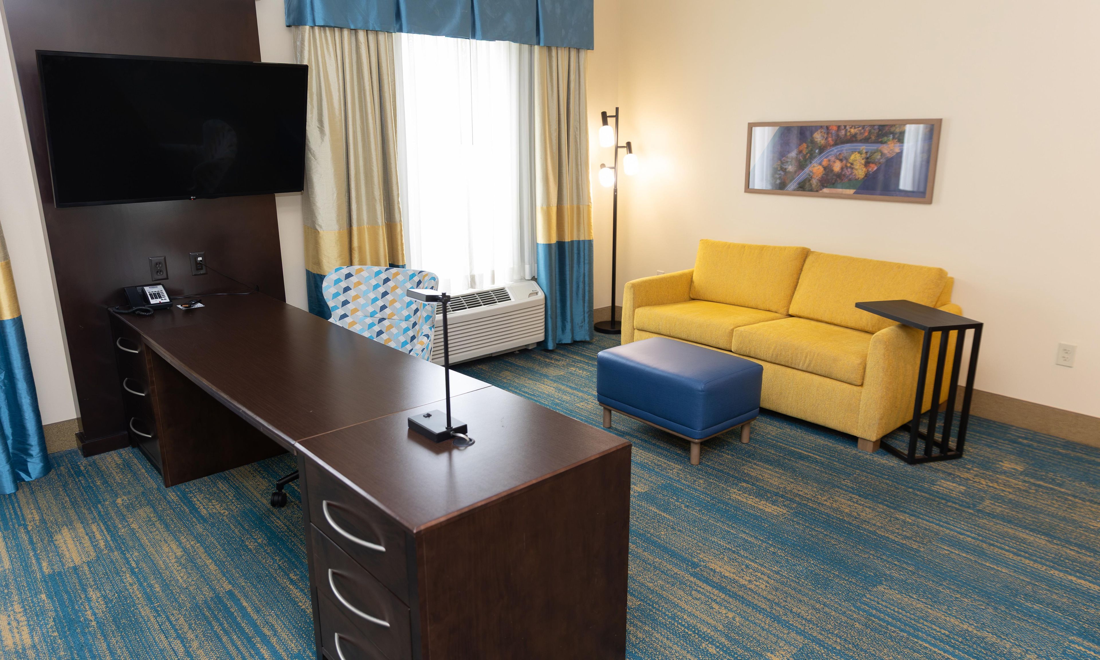 A hotel room with a desk, a sofa, a TV, a painting, and a lamp.