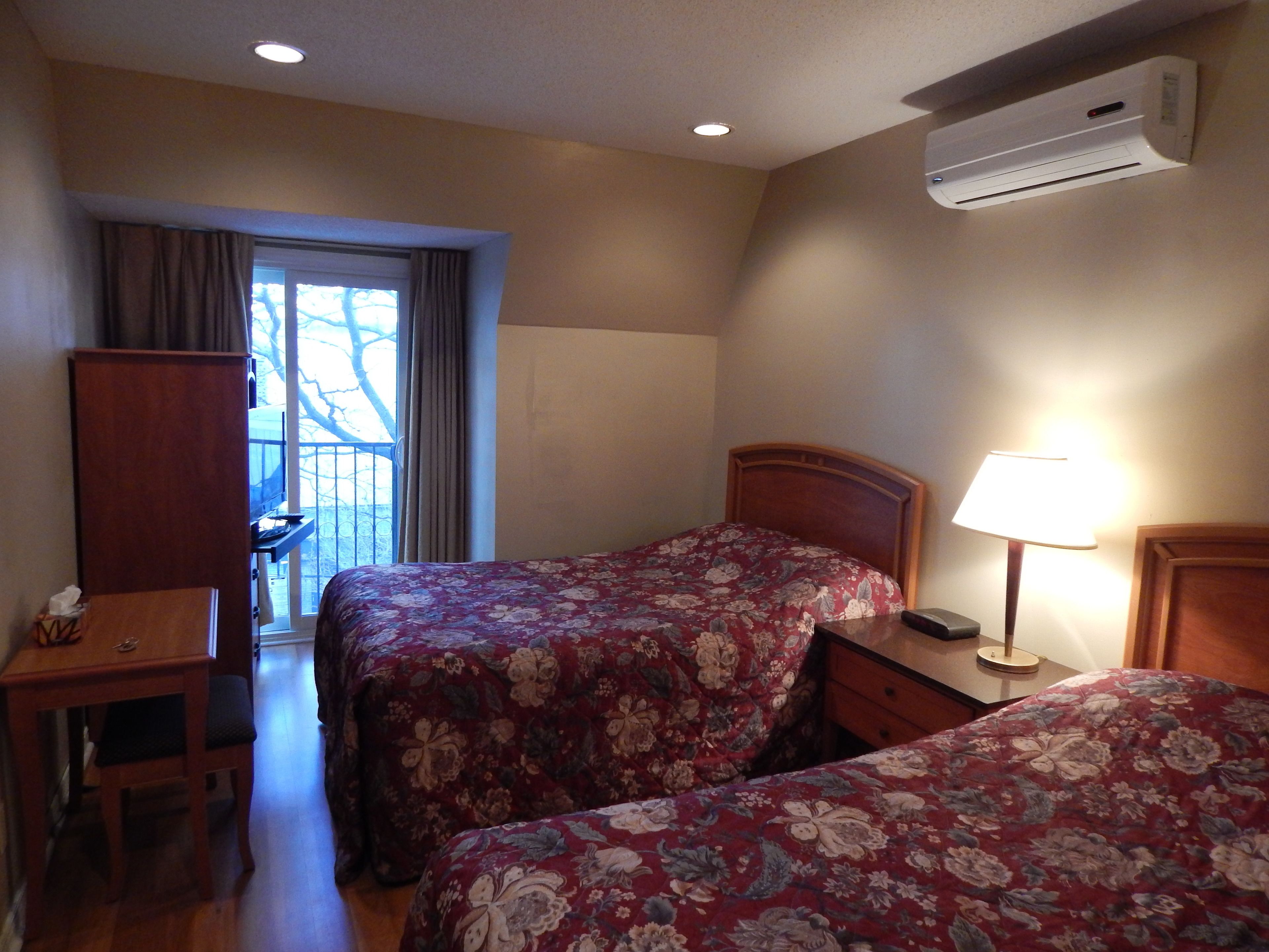 A hotel room with two beds, a desk, a chair, a lamp, and an air conditioning unit.