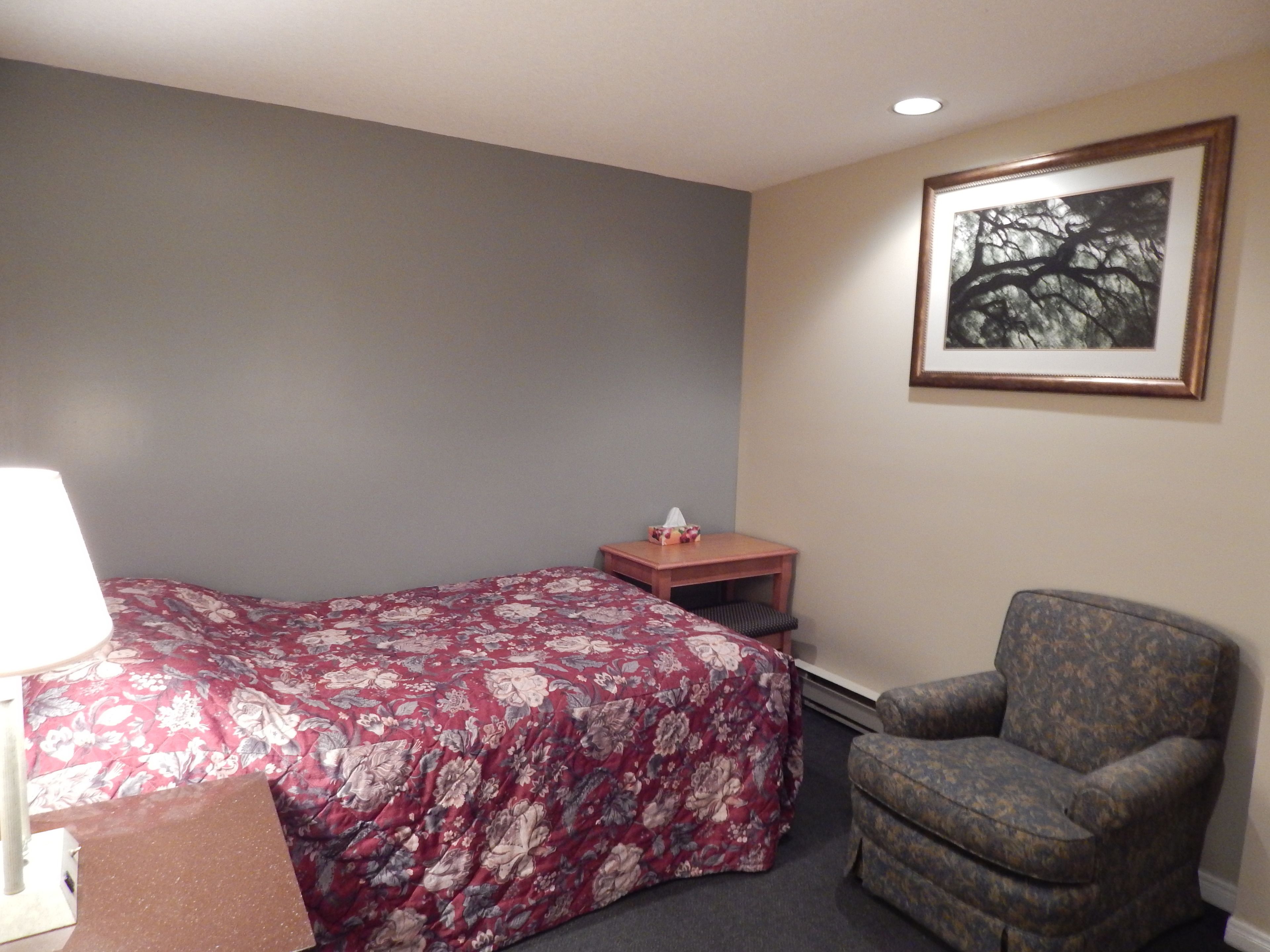 Quarto de hotel com cama, uma cadeira, uma mesinha com um porta-lenços e um quadro na parede.