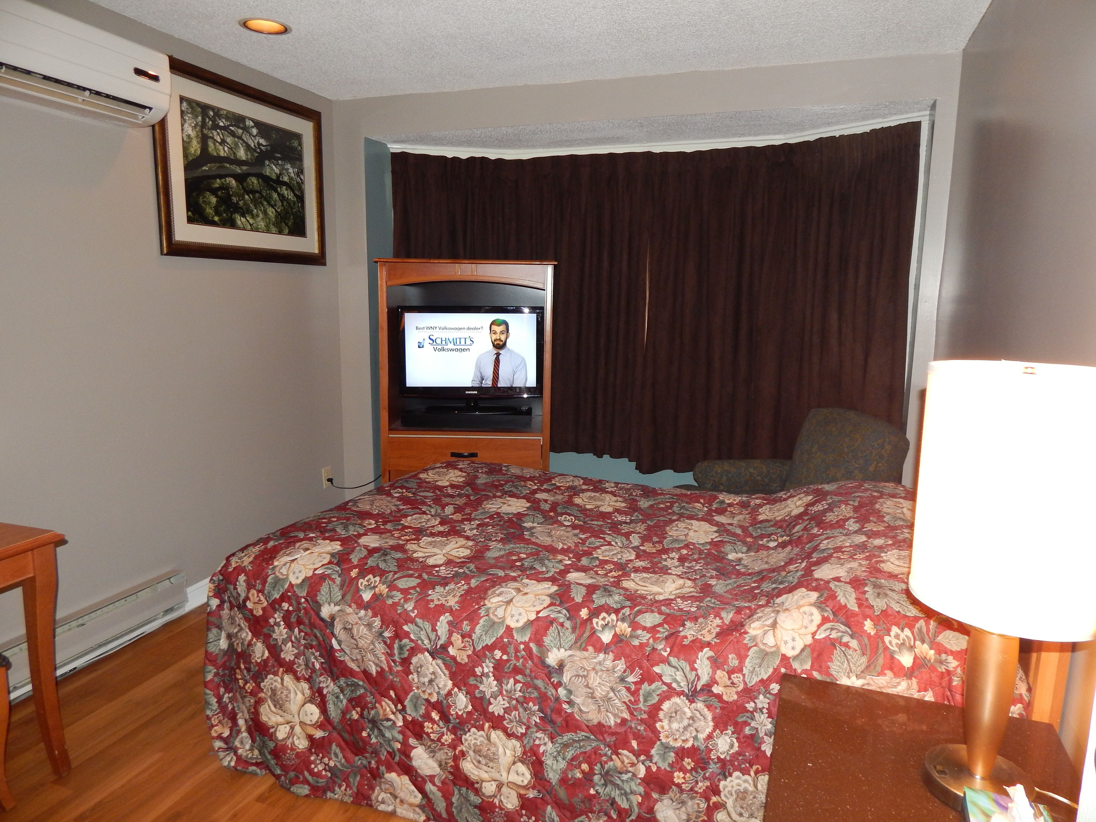 A hotel room with a bed, a television, a window with curtains, and a framed picture on the wall.