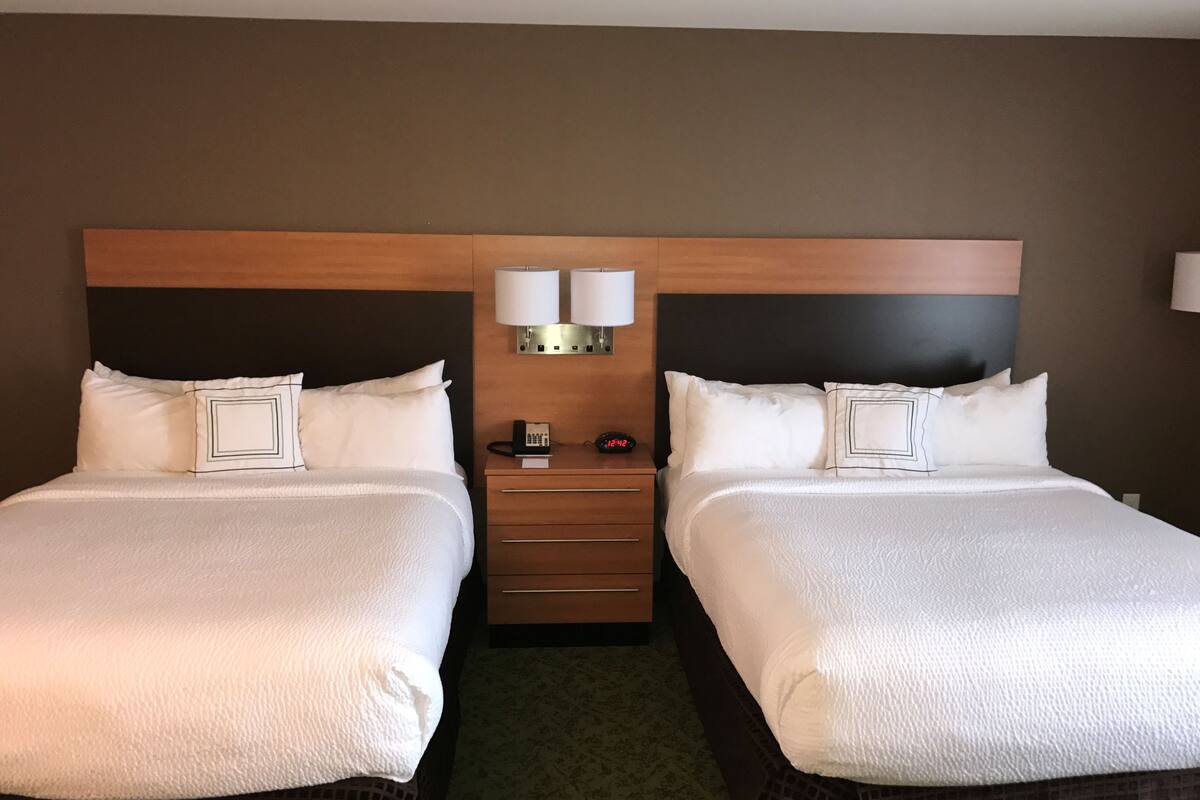 A hotel room with two beds, a nightstand with a phone and a clock, and a wall-mounted lamp.