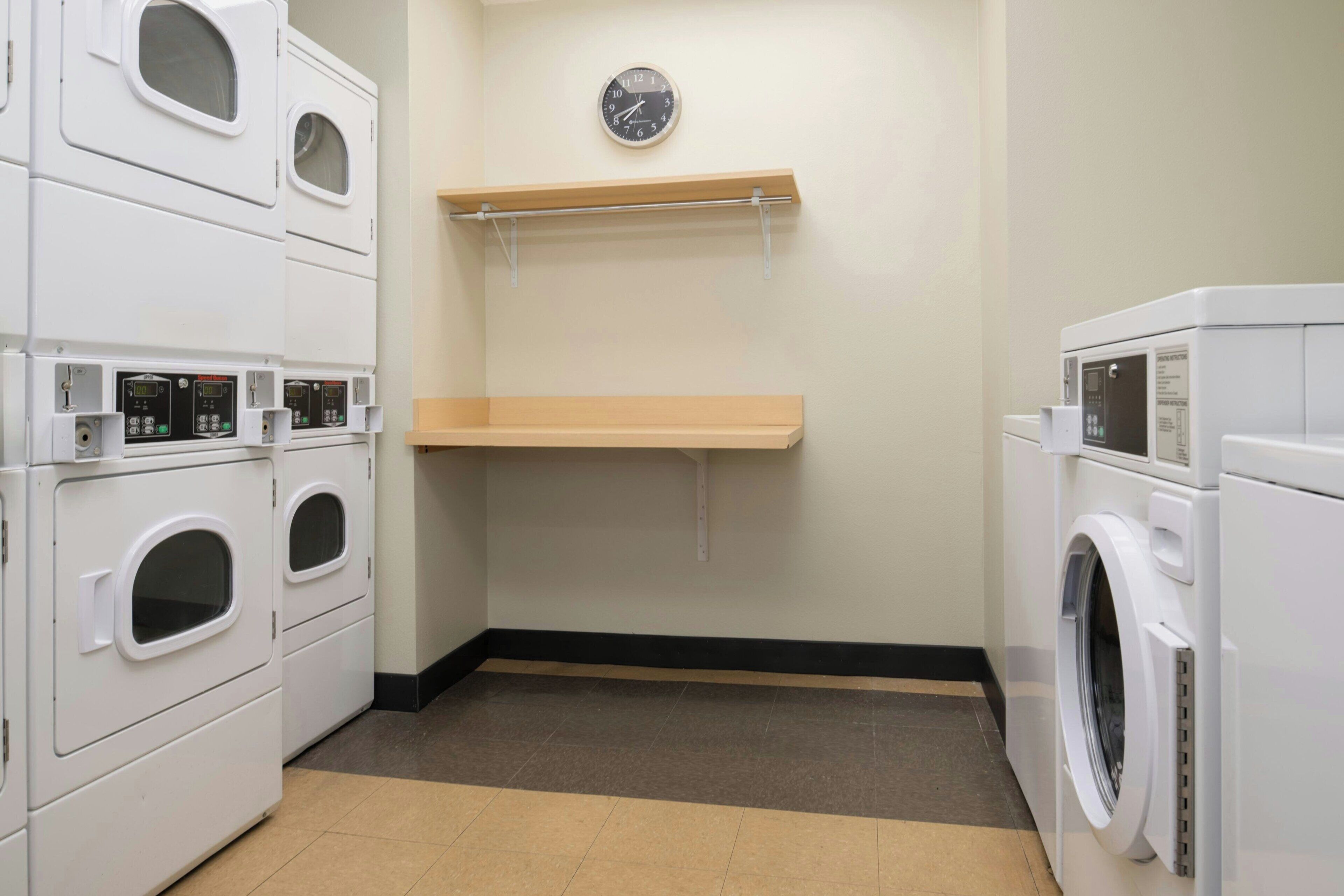 A laundry room with stacked washers and dryers, a clock on the wall, and a shelf above.