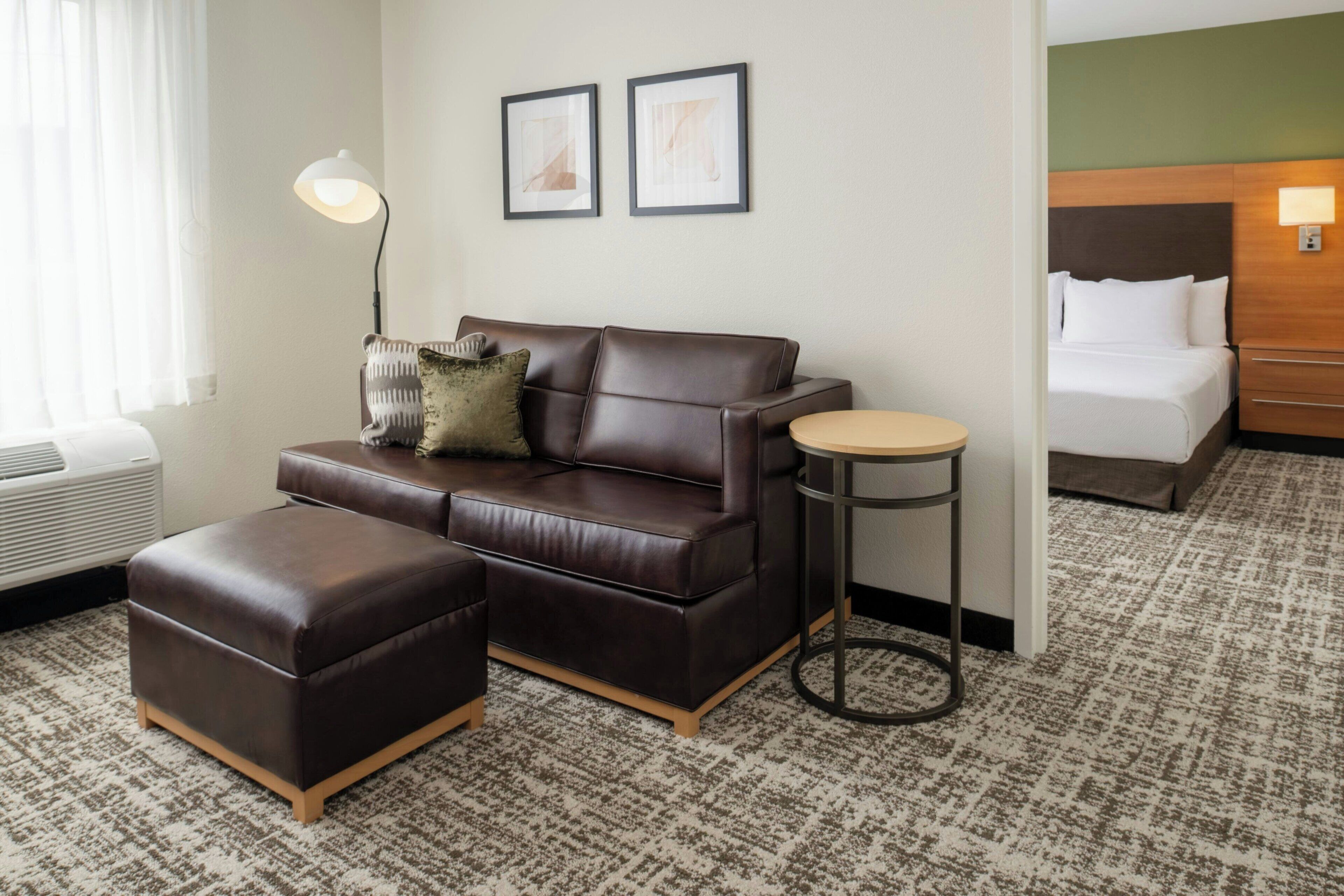 A hotel room with a brown leather sofa, a matching ottoman, a round side table, a bed with white linens, and two framed pictures on the wall.