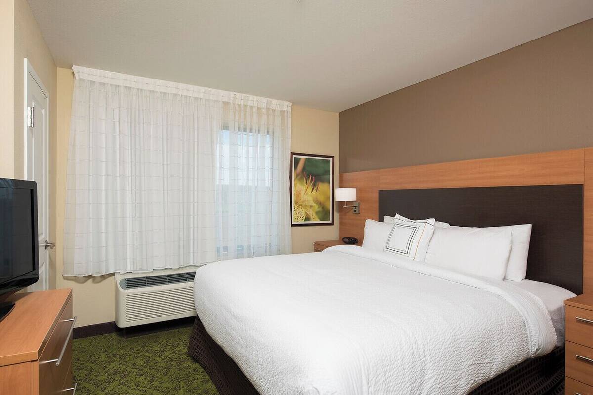 A hotel room with a large bed, a television, a heater, a window with curtains, and a painting on the wall.