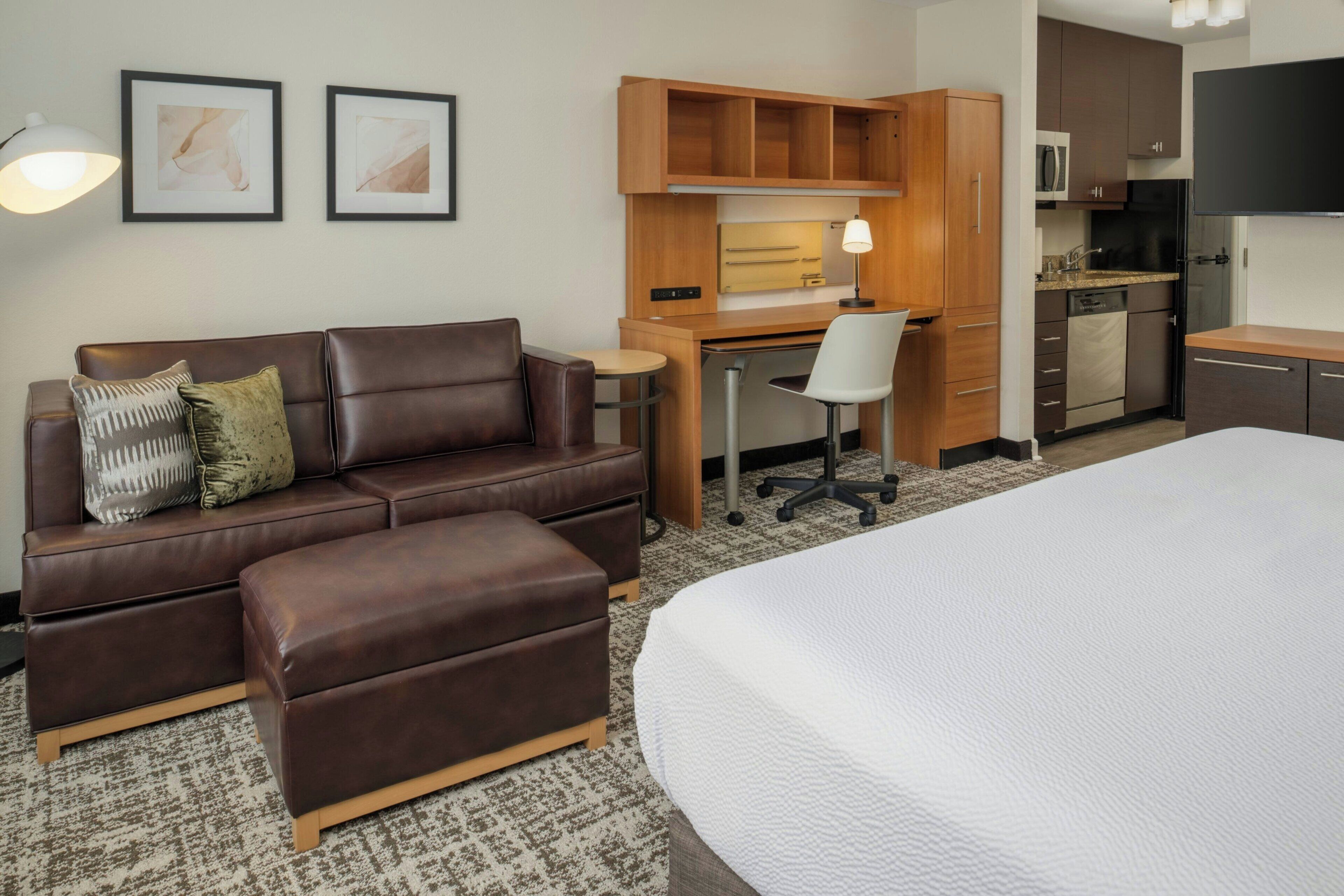 A hotel room with a brown leather sofa, a small desk with a chair, a bed, a kitchenette, and two framed pictures on the wall.