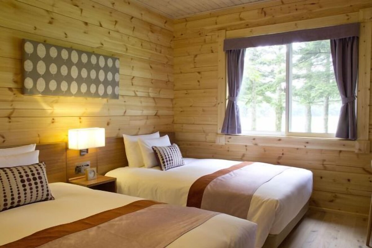 A wooden cabin room with two beds, a nightstand with a lamp, and a window with curtains.