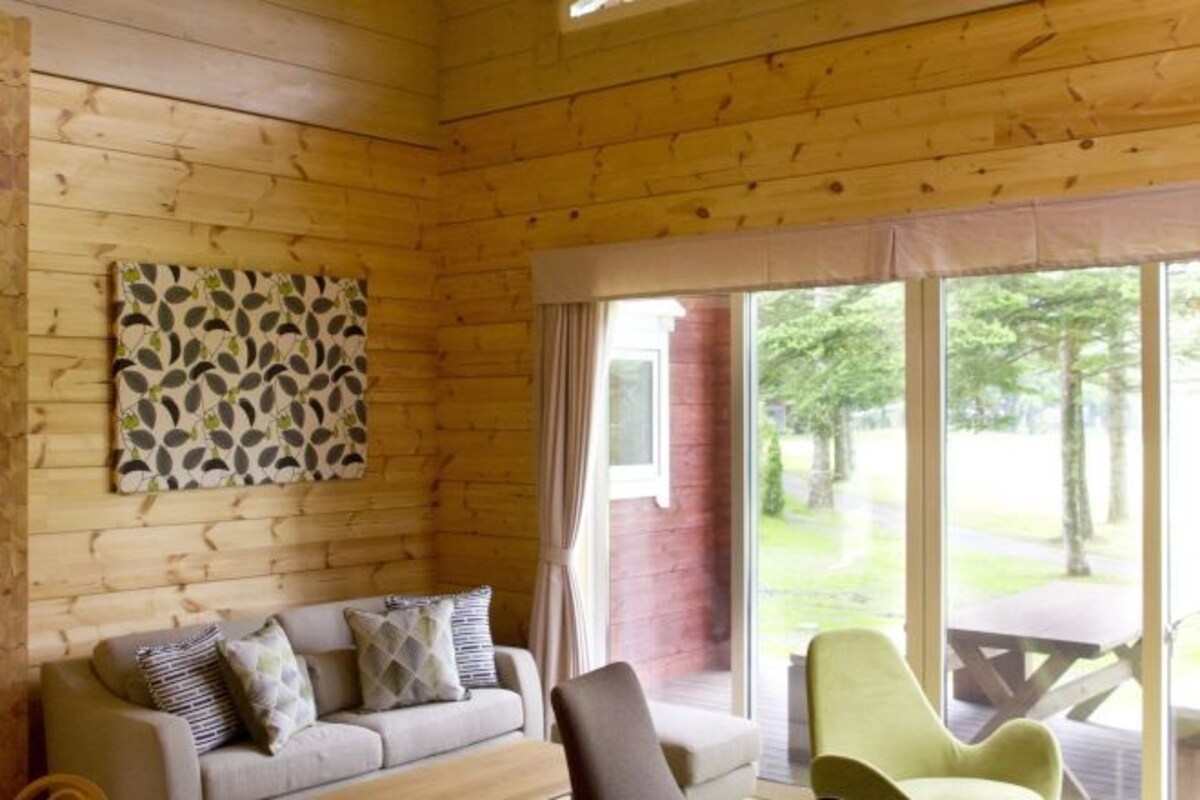 Interior de una cabaña de madera con un sofá, una silla verde y un ventanal con vistas al exterior.