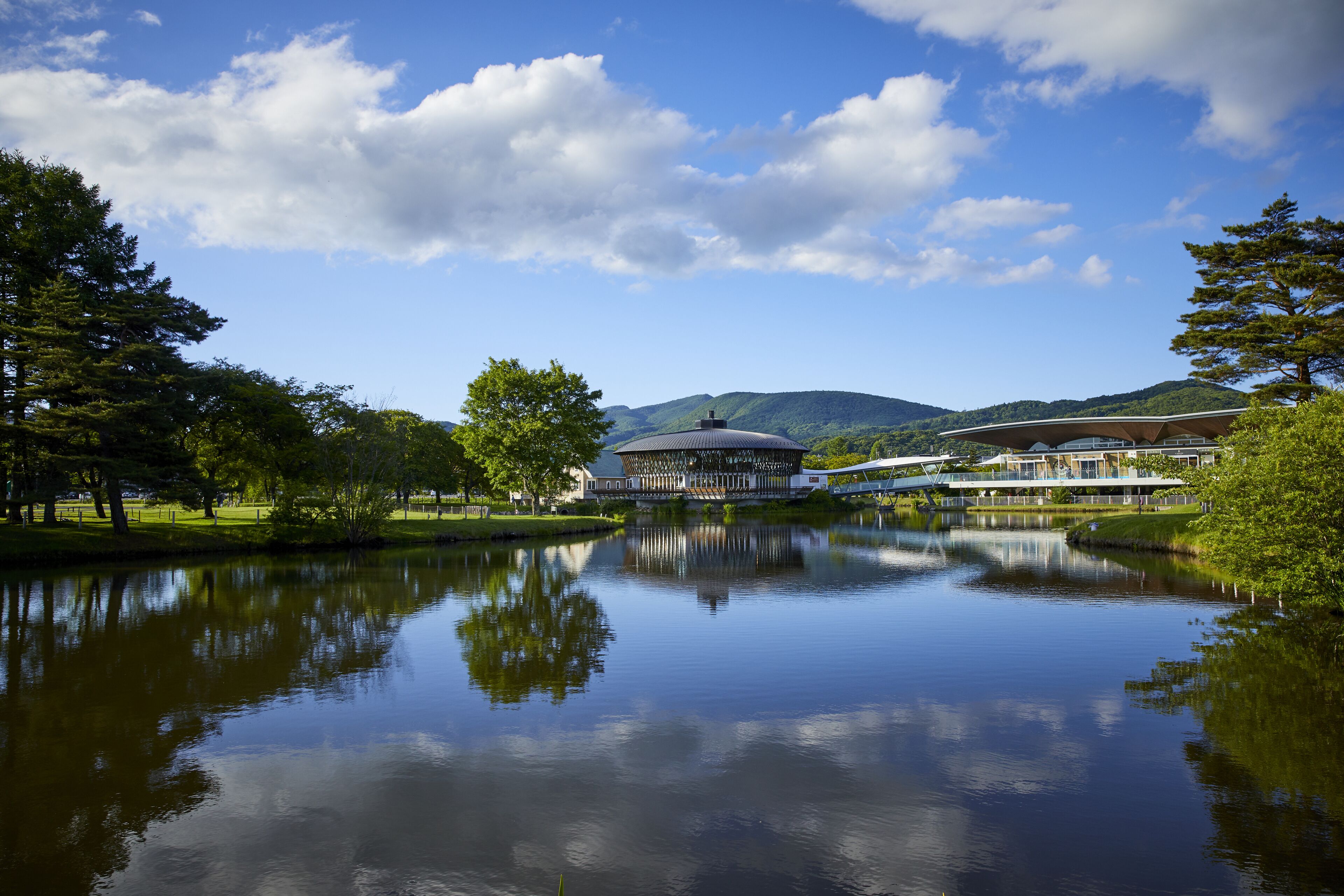A serene lake with a reflection of a building and trees, surrounded by a park and mountains in the background.