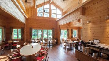 A spacious wooden cabin with multiple dining tables and chairs, large windows, and a kitchen area.