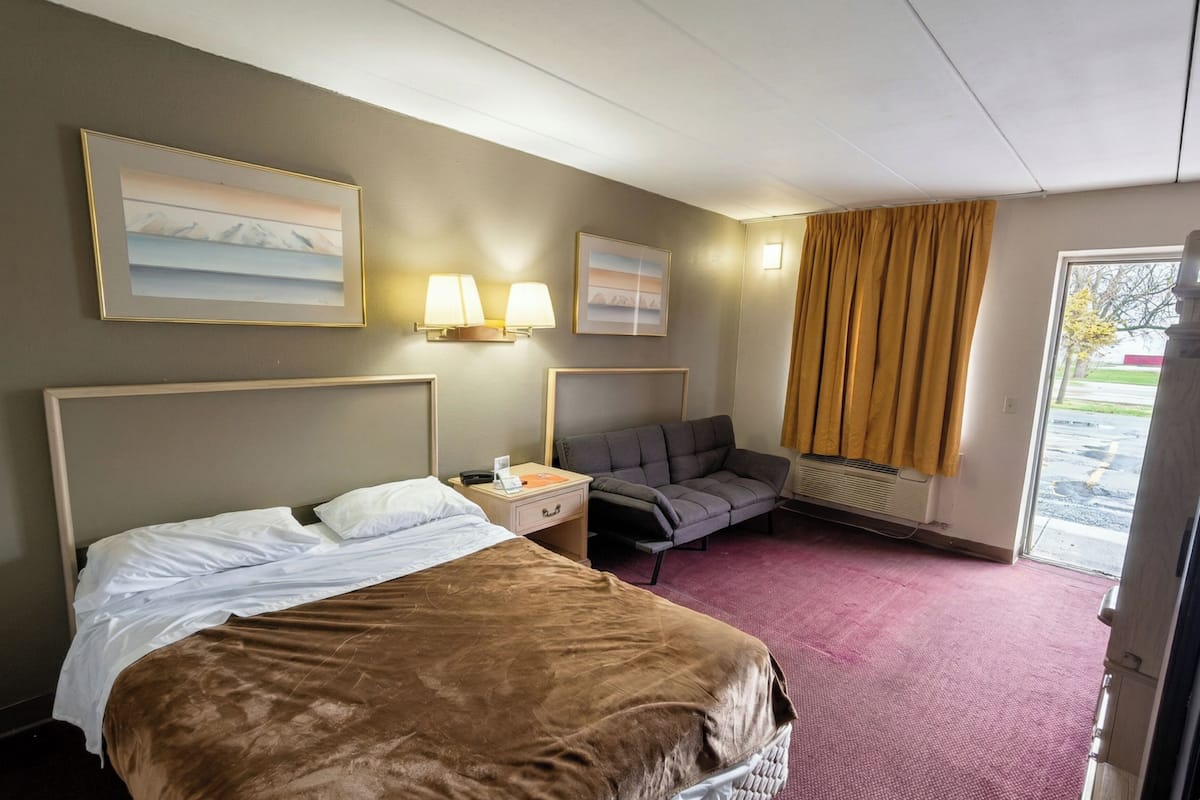 Ein Hotelzimmer mit einem Bett, einem Schreibtisch, einem Stuhl, einem Fenster mit Vorhängen und Bildern an den Wänden.