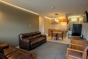 A living room with a brown leather sofa, a dining table with chairs, a kitchen area with wooden cabinets, and a flat-screen TV mounted on the wall. - Lake Erie Lodge Oregon OH (Curtice)