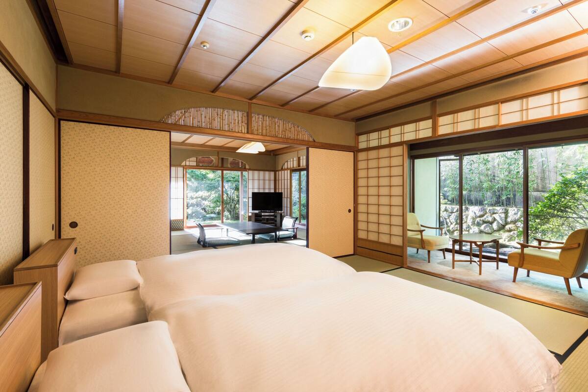 A traditional Japanese-style bedroom with tatami flooring, a large bed, a television, and a view of a garden through large windows.