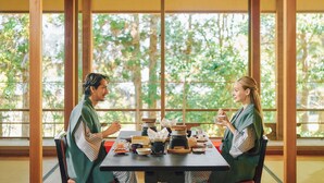 Una habitación japonesa tradicional con suelo de tatami, una mesa baja con utensilios para la ceremonia del té y grandes ventanales que dan a vistas de follaje exterior.