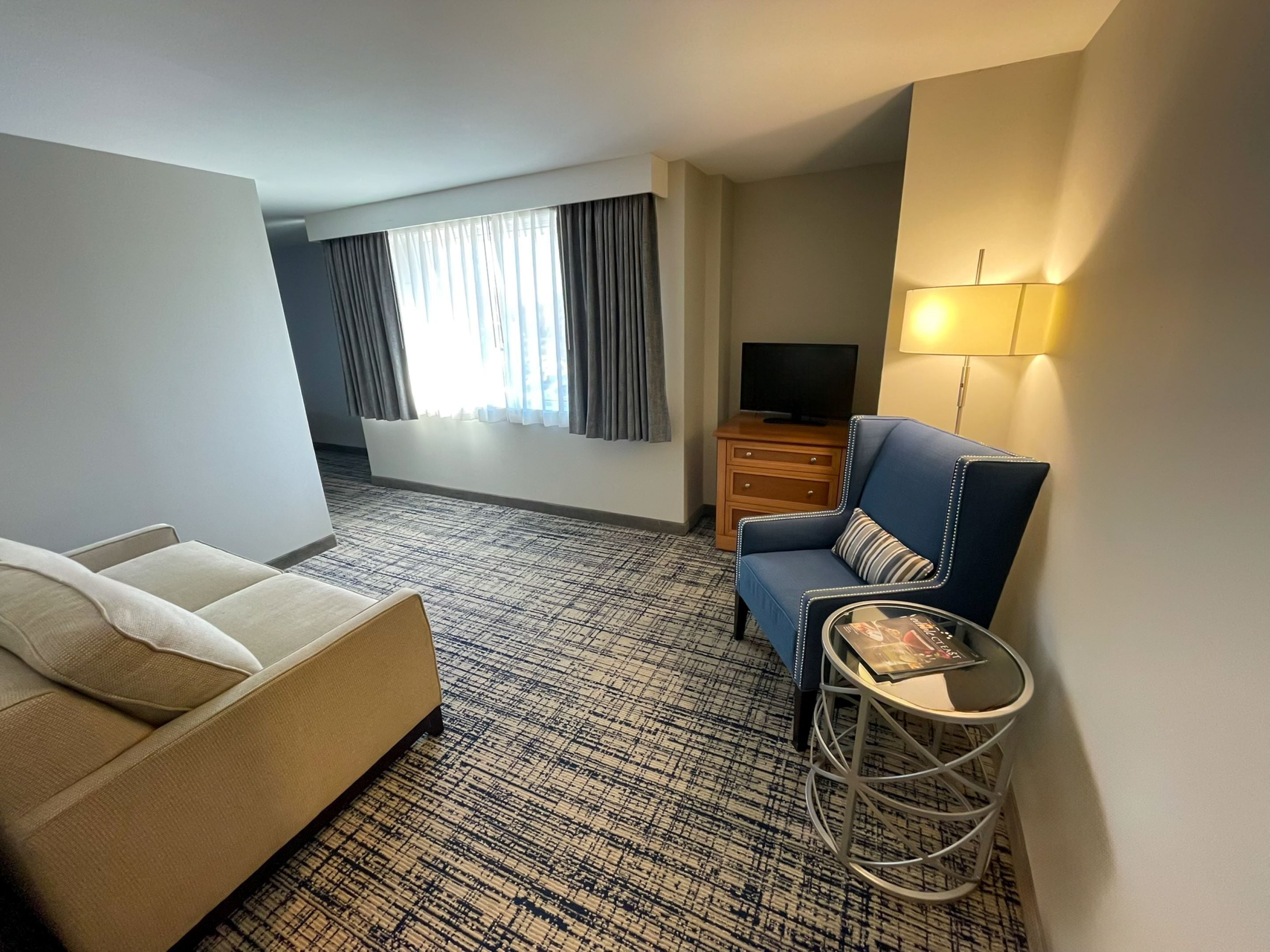Habitación de hotel con sofá, una silla, una mesita, televisión y lámpara.