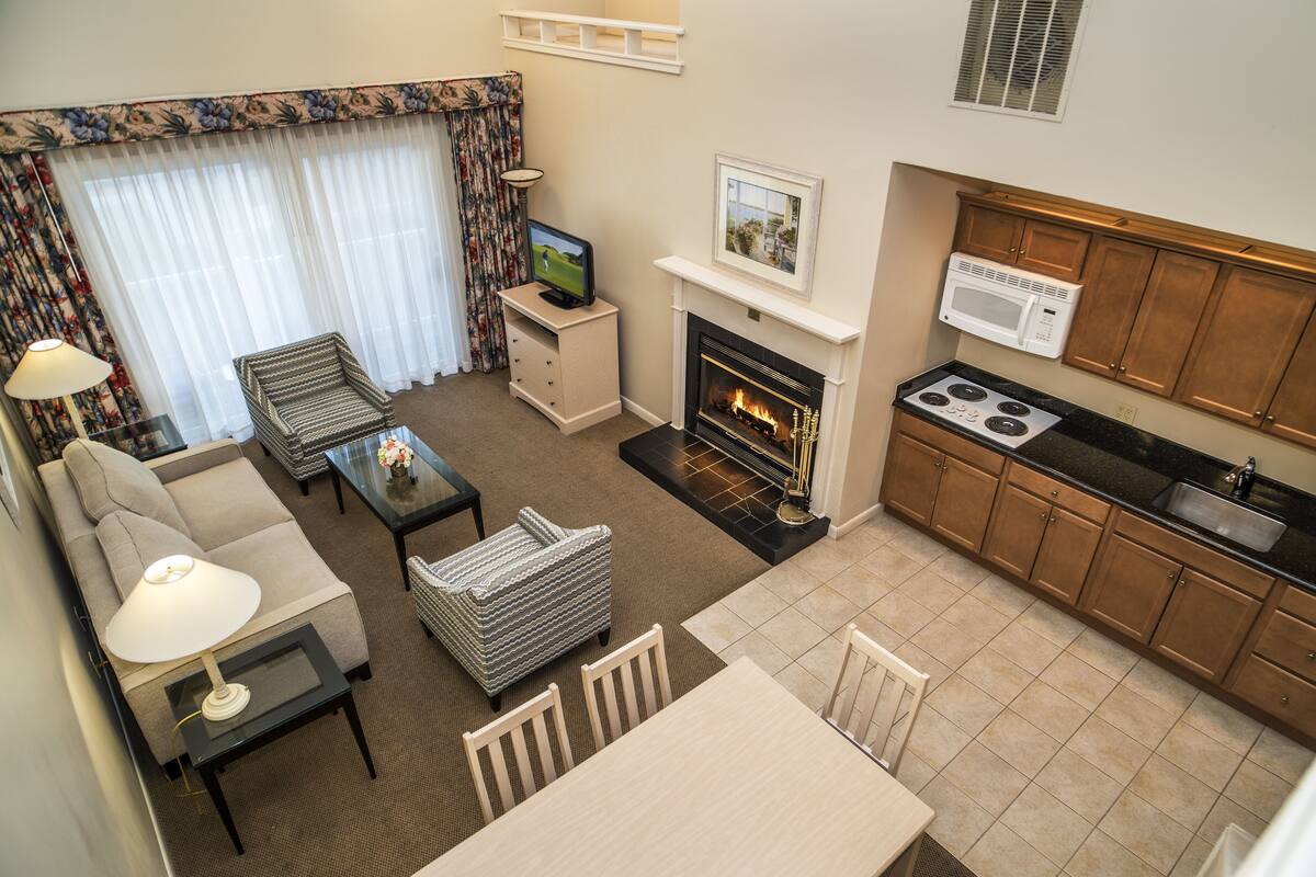 A compact living space with a sofa, armchair, and a small dining table. A fireplace is centrally located, and a kitchen area with a stove and microwave is visible.