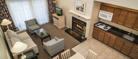 A compact living space with a sofa, armchair, and a small dining table. A fireplace is centrally located, and a kitchen area with a stove and microwave is visible.