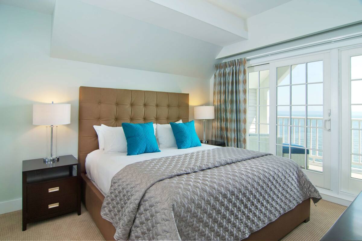 A hotel room with a large bed, two bedside lamps, a nightstand, and a view of the sea through a sliding glass door.