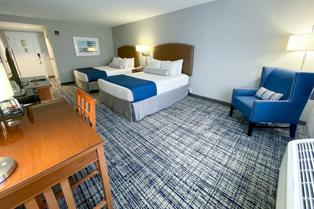 A hotel room with two beds, a desk, a chair, and a patterned carpet.