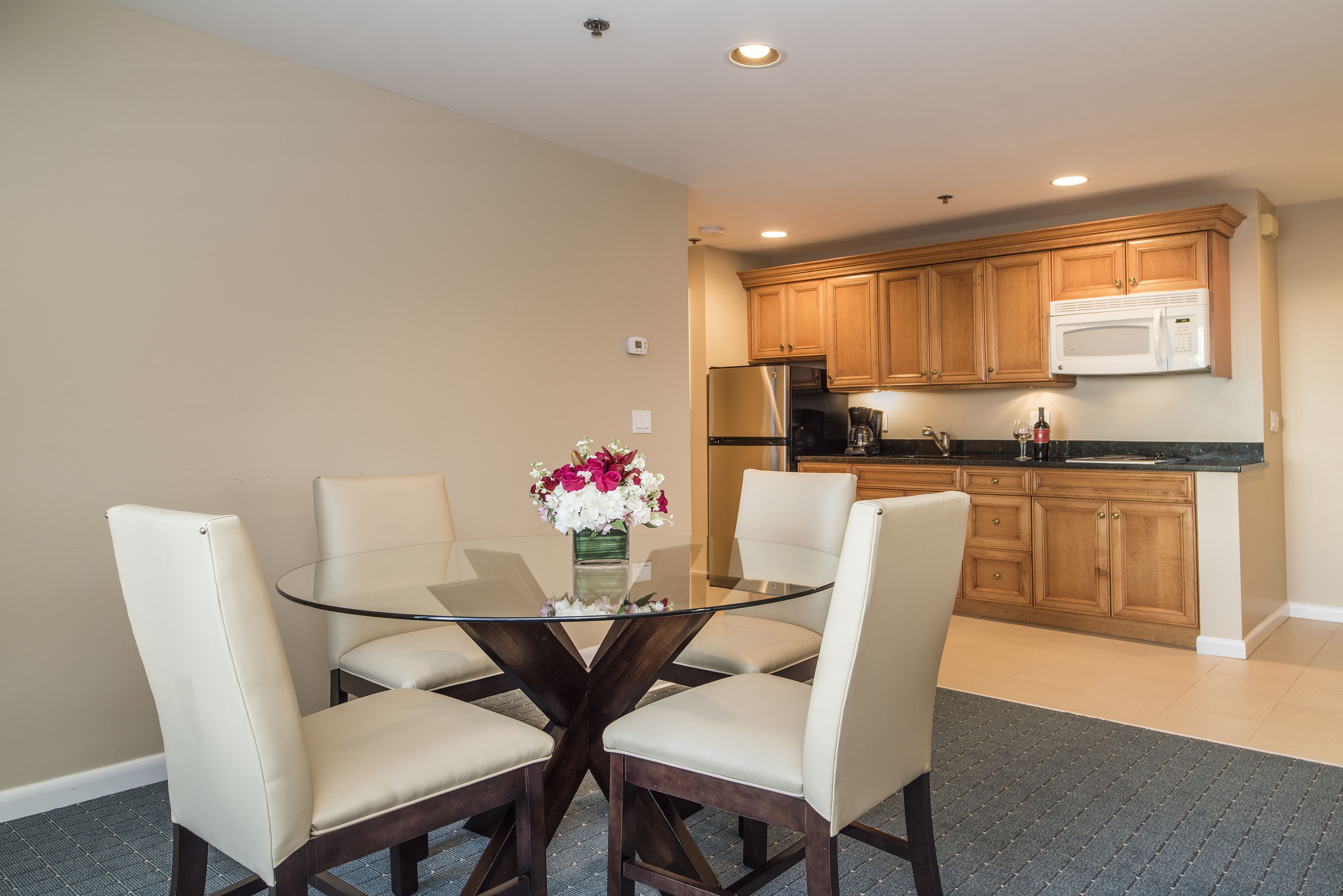 A dining area with a glass table surrounded by four chairs, a vase with flowers, and a kitchen in the background with wooden cabinets and appliances.