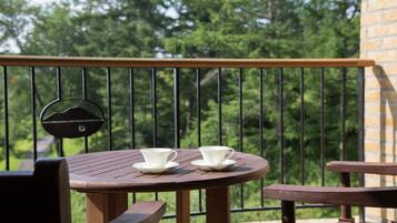 A wooden table with two white cups on saucers, a black chair, and a metal railing with a view of trees and a brick wall in the background.