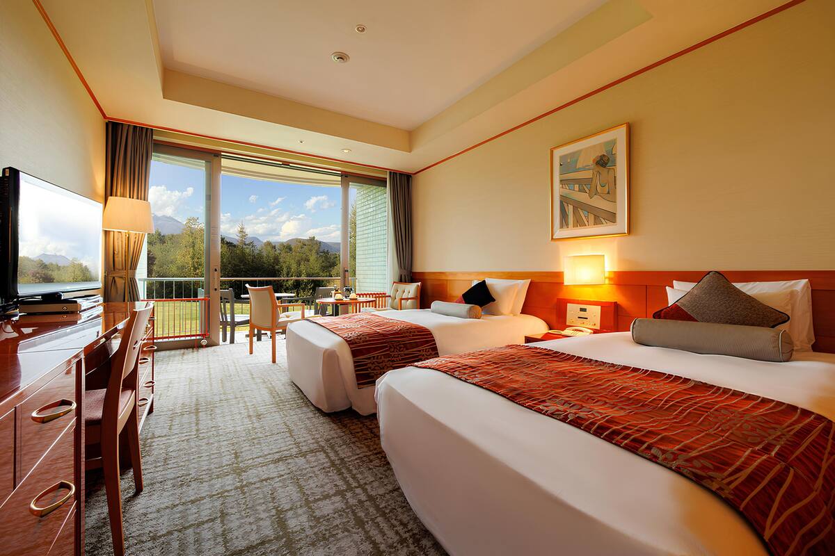 A hotel room with two beds, a desk, a TV, and a balcony with a view of mountains.