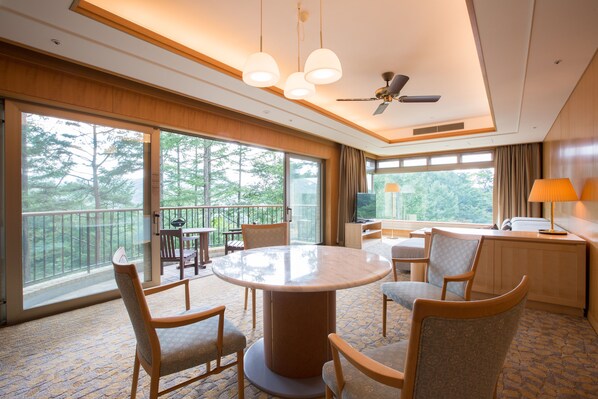 Habitación de hotel con un ventanal grande, una mesa redonda, varias sillas, un ventilador de techo y un televisor.
