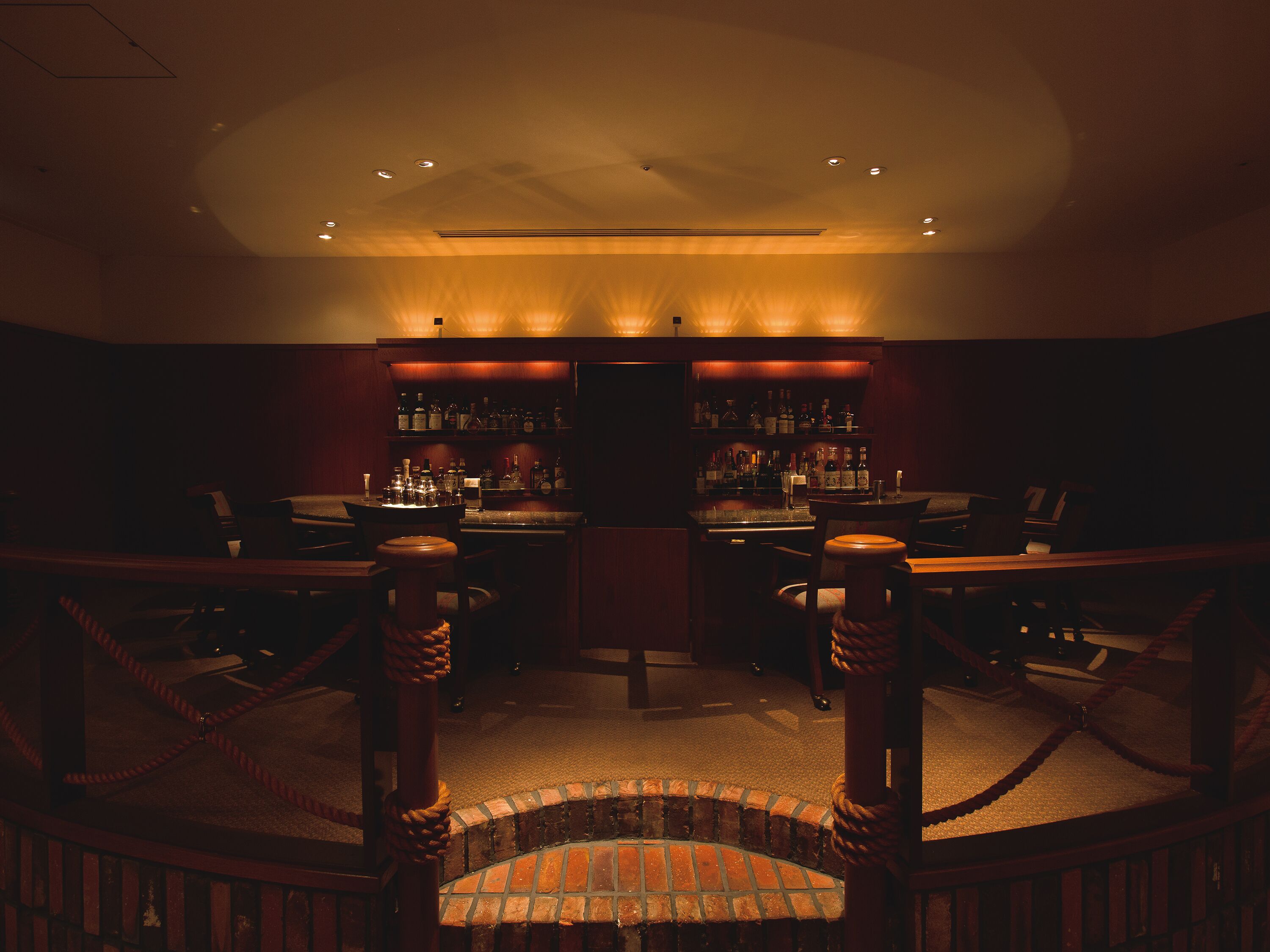 Zona de bar con poca luz, chimenea de ladrillo, taburetes de madera y una variedad de botellas en estanterías.