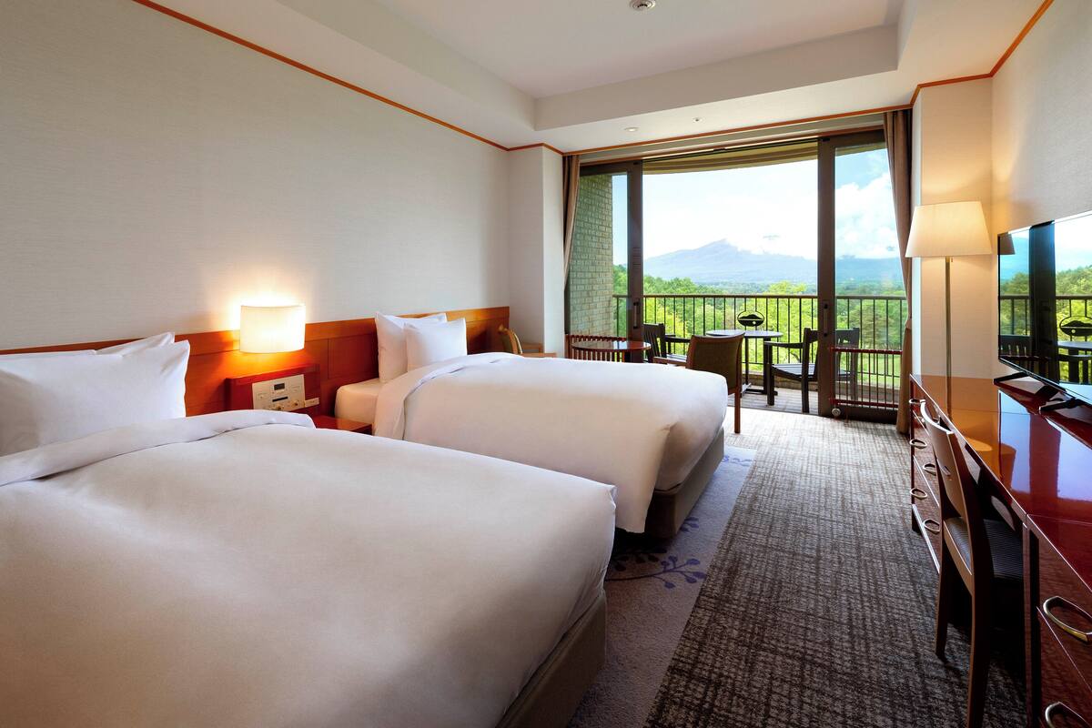 A hotel room with two beds, a desk, and a balcony with a view.