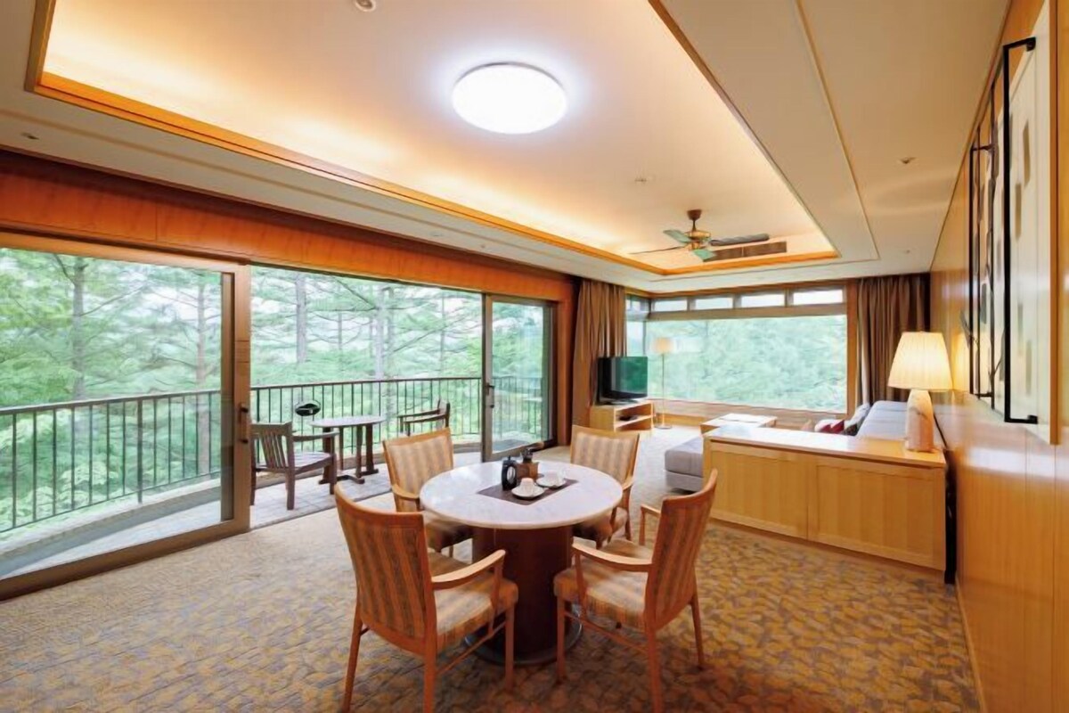 Habitación de hotel con balcón, una mesa redonda de comedor con sillas, un sofá, un televisor y una lámpara.