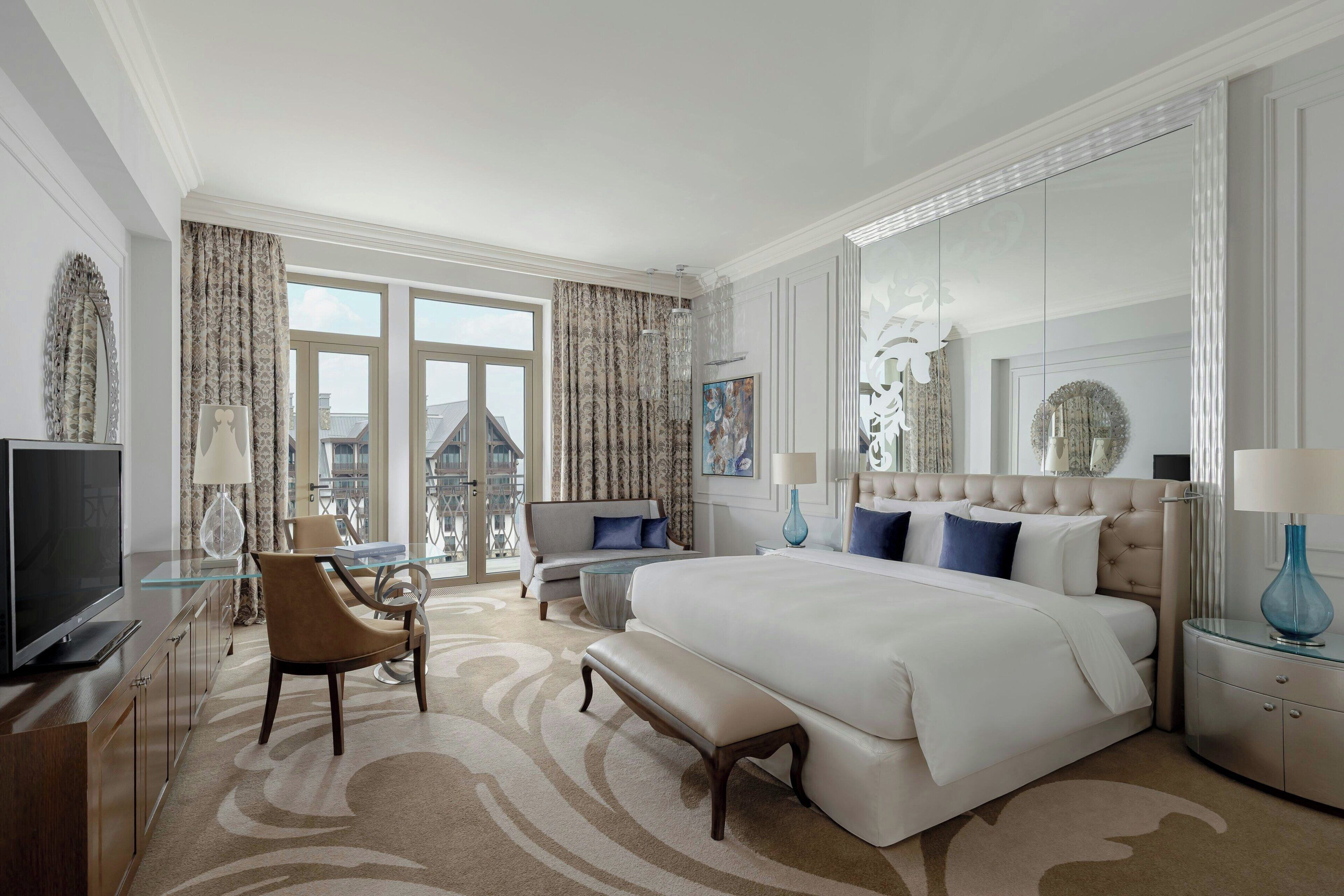 A spacious hotel room with a large bed, a seating area with a sofa and armchairs, a television, and a view of the city through large windows.