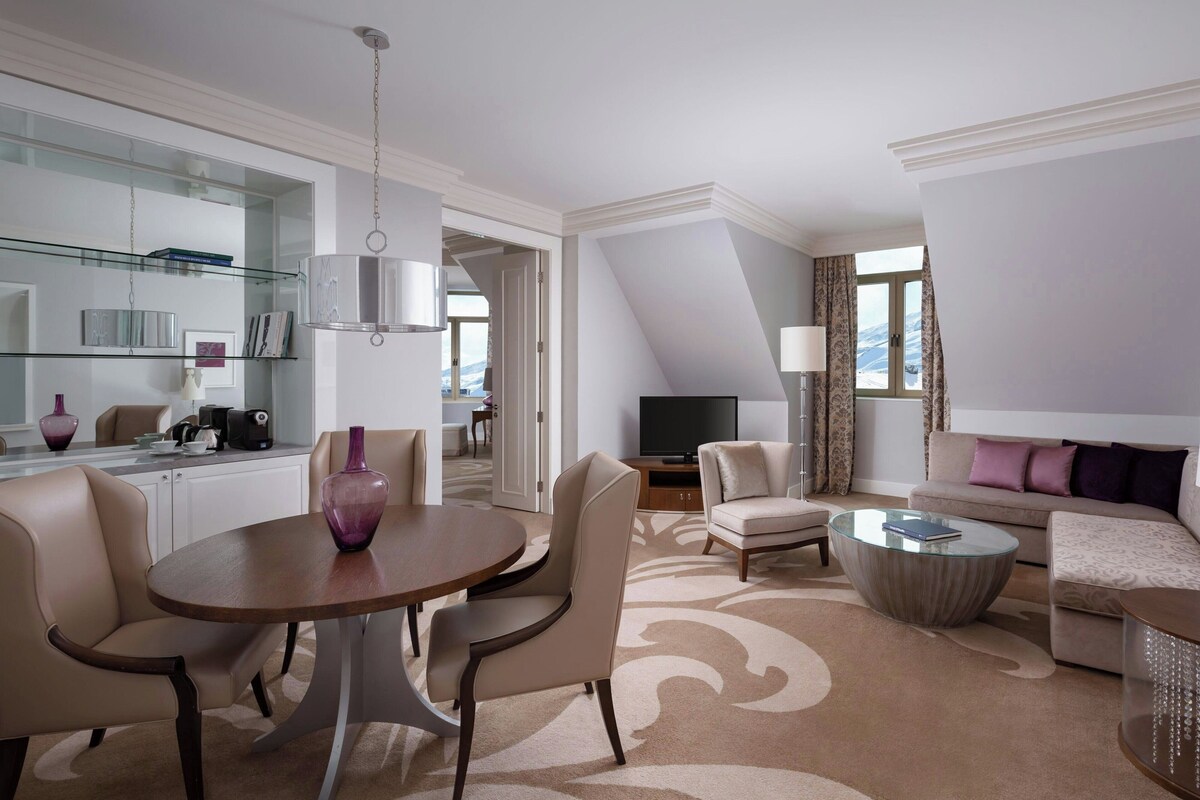 Una habitación de hotel moderna con zona de comedor, sala de estar con sofá y sillones, televisor y un amplio ventanal con cortinas.