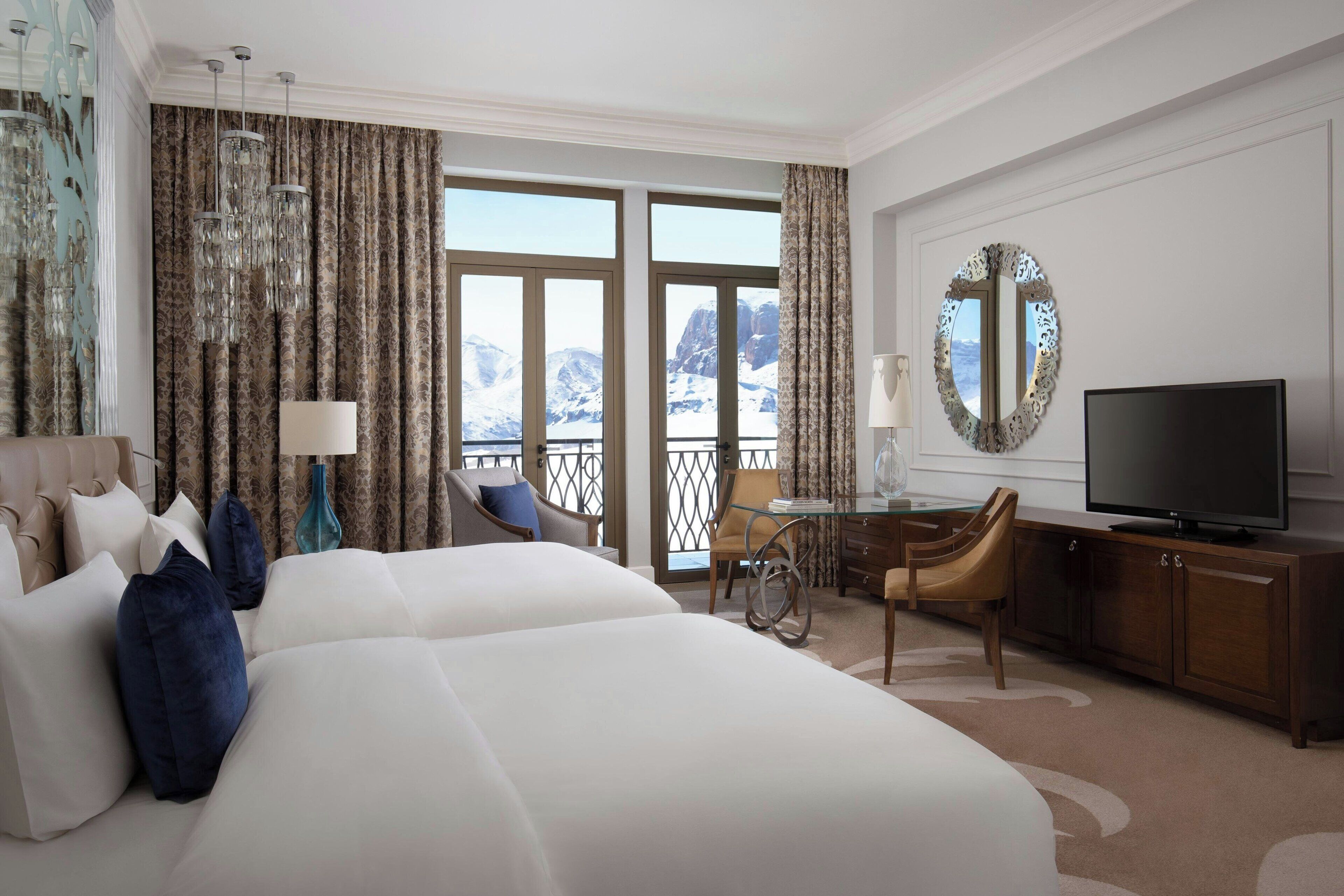 A hotel room with a large bed, a desk, a television, and a view of mountains through the windows.
