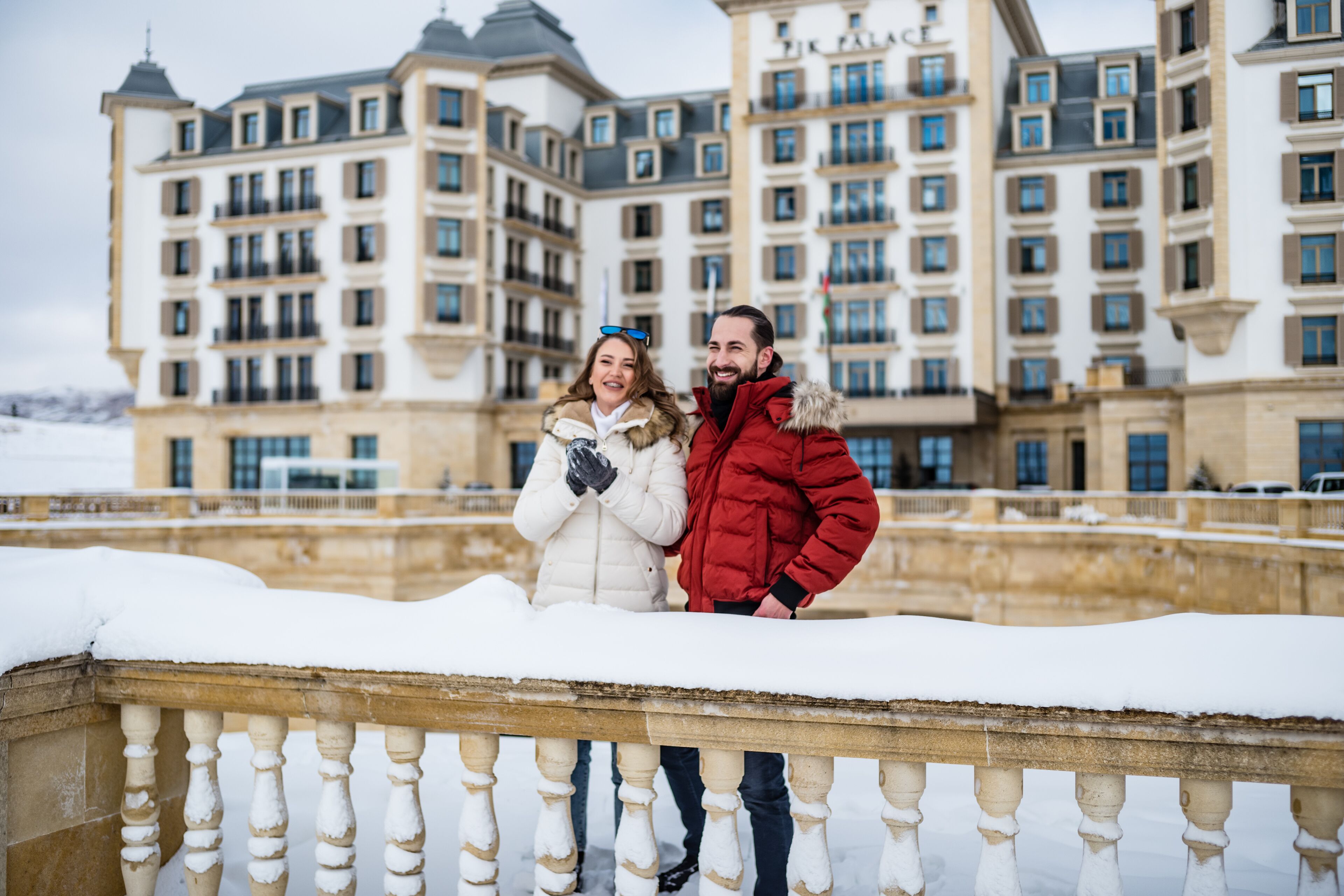 A couple standing on a snow-covered balcony with a large, historic building in the background.