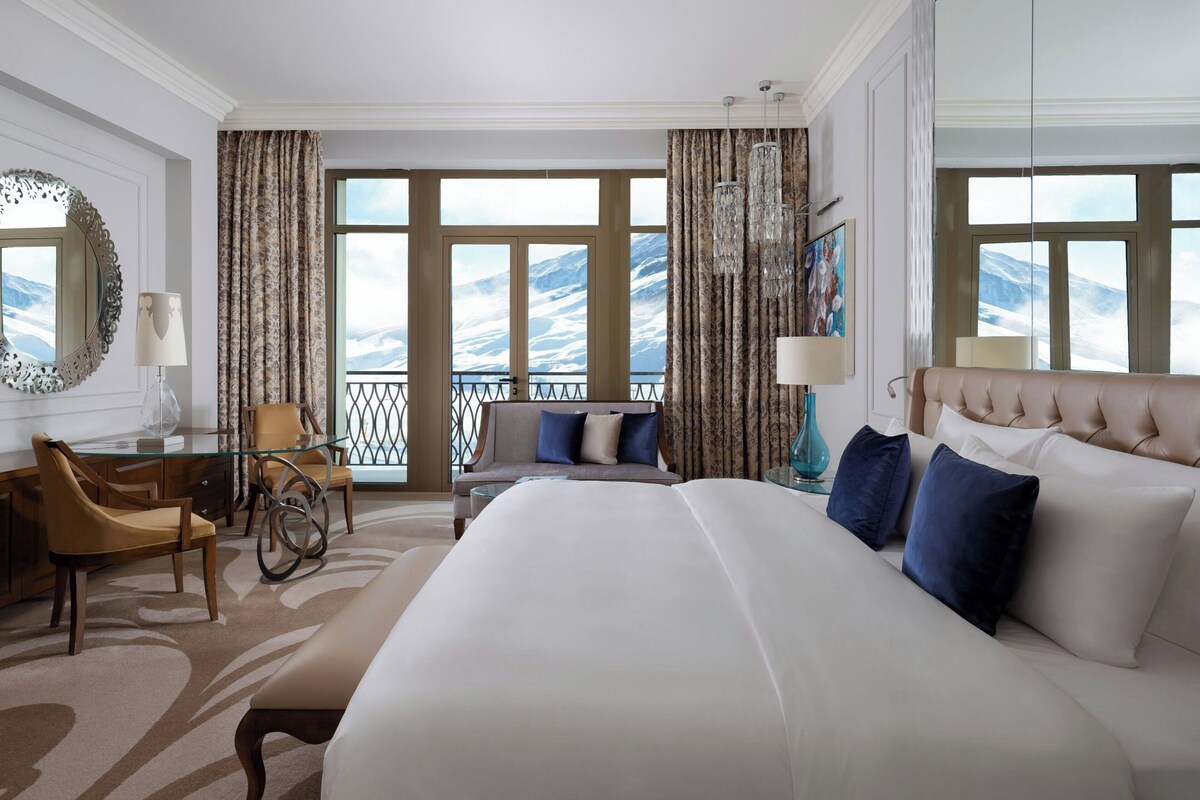 Habitación de hotel con una cama grande, un escritorio con una silla, un sofá y vistas a montañas a través de amplios ventanales.