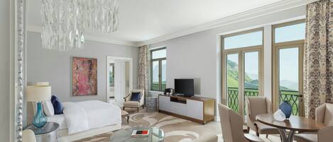 A hotel room with a bed, a TV, a dining table with chairs, a sofa, a coffee table, and a balcony with a view of the hills.
