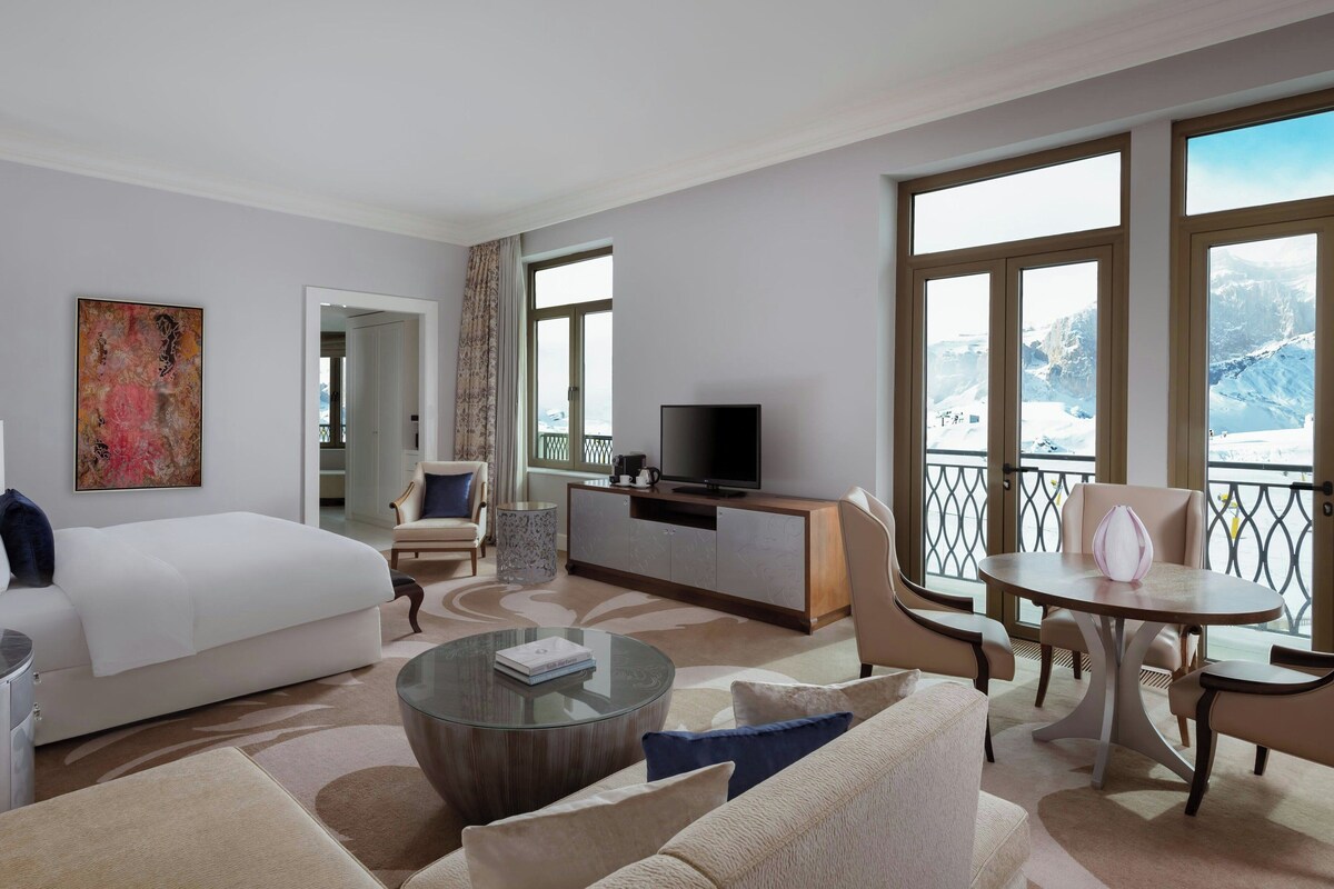 Habitación de hotel moderna con una cama grande, televisión, zona de comedor y balcón con vistas a la montaña.