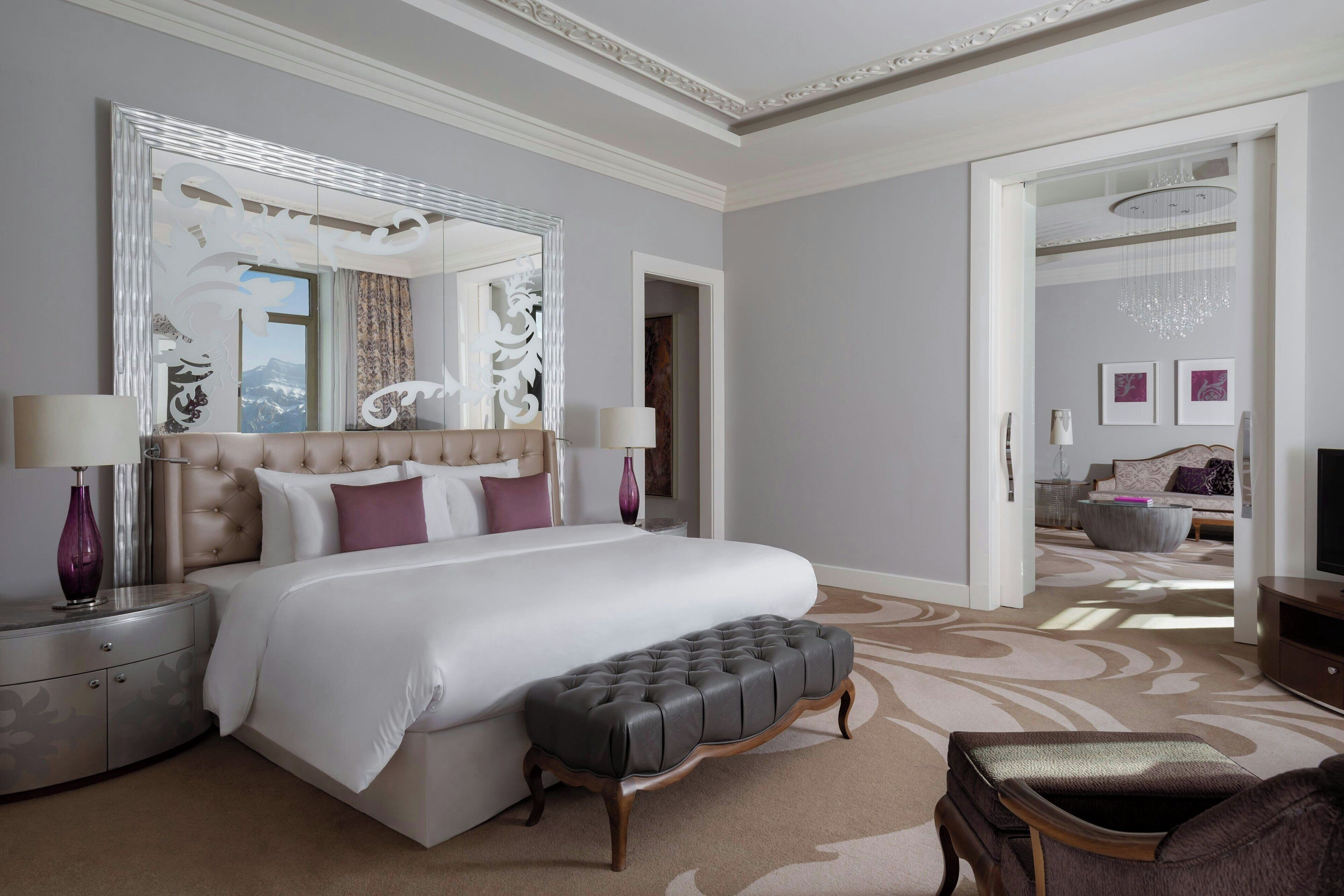 A spacious bedroom with a large bed, a tufted bench, a nightstand, and a mirror.