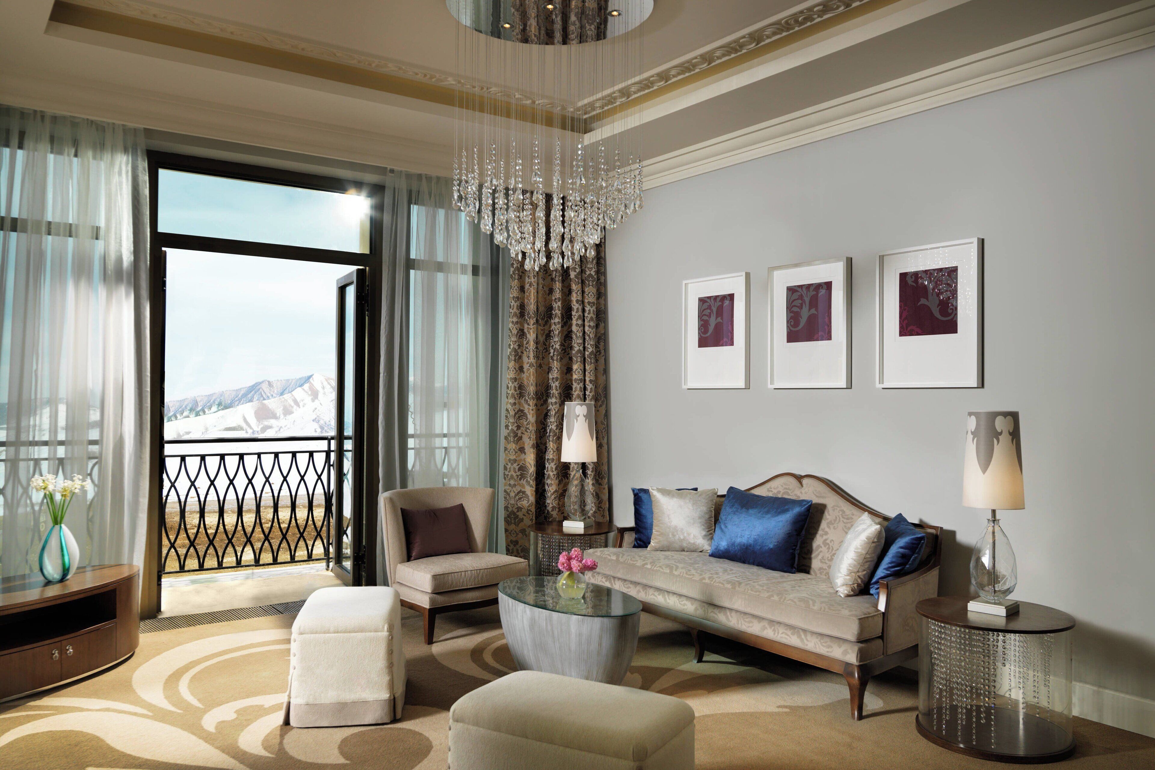 A spacious living room with a sofa, armchairs, a coffee table, and a chandelier. There are three framed pictures on the wall and a view through a large window.